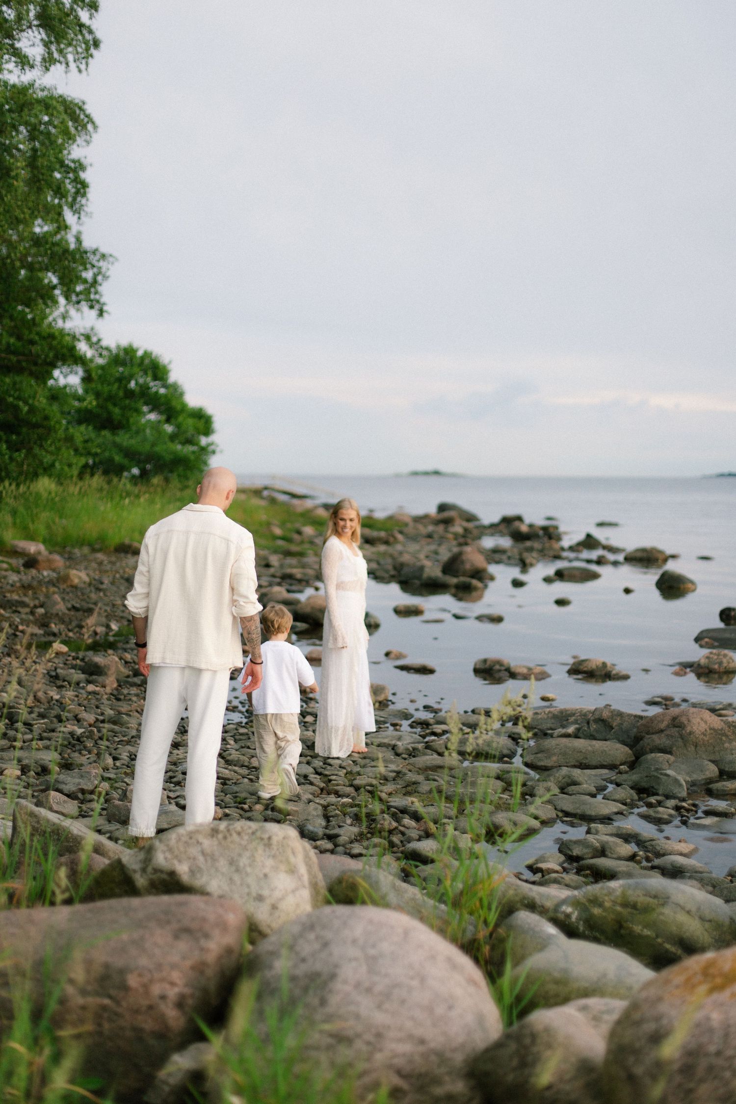 Family in white clothing walking along a rocky shoreline near lush green trees and calm ocean waters.