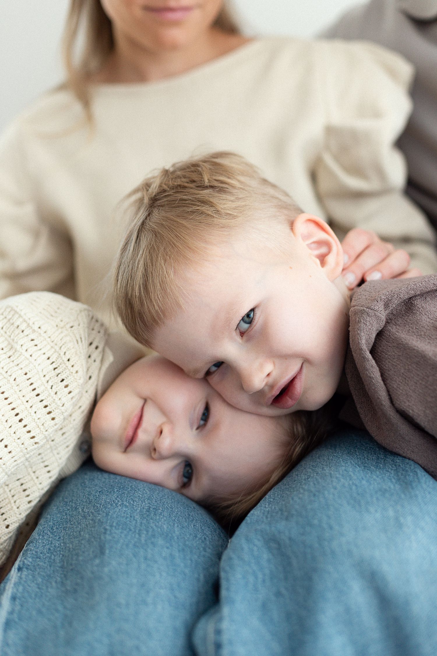 Two young siblings lean together on their mother's lap, smiling warmly in a cozy family portrait.