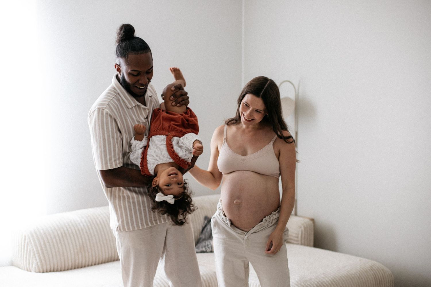 A happy interracial family with a pregnant woman and man holding a baby and toddler in a bright white room.