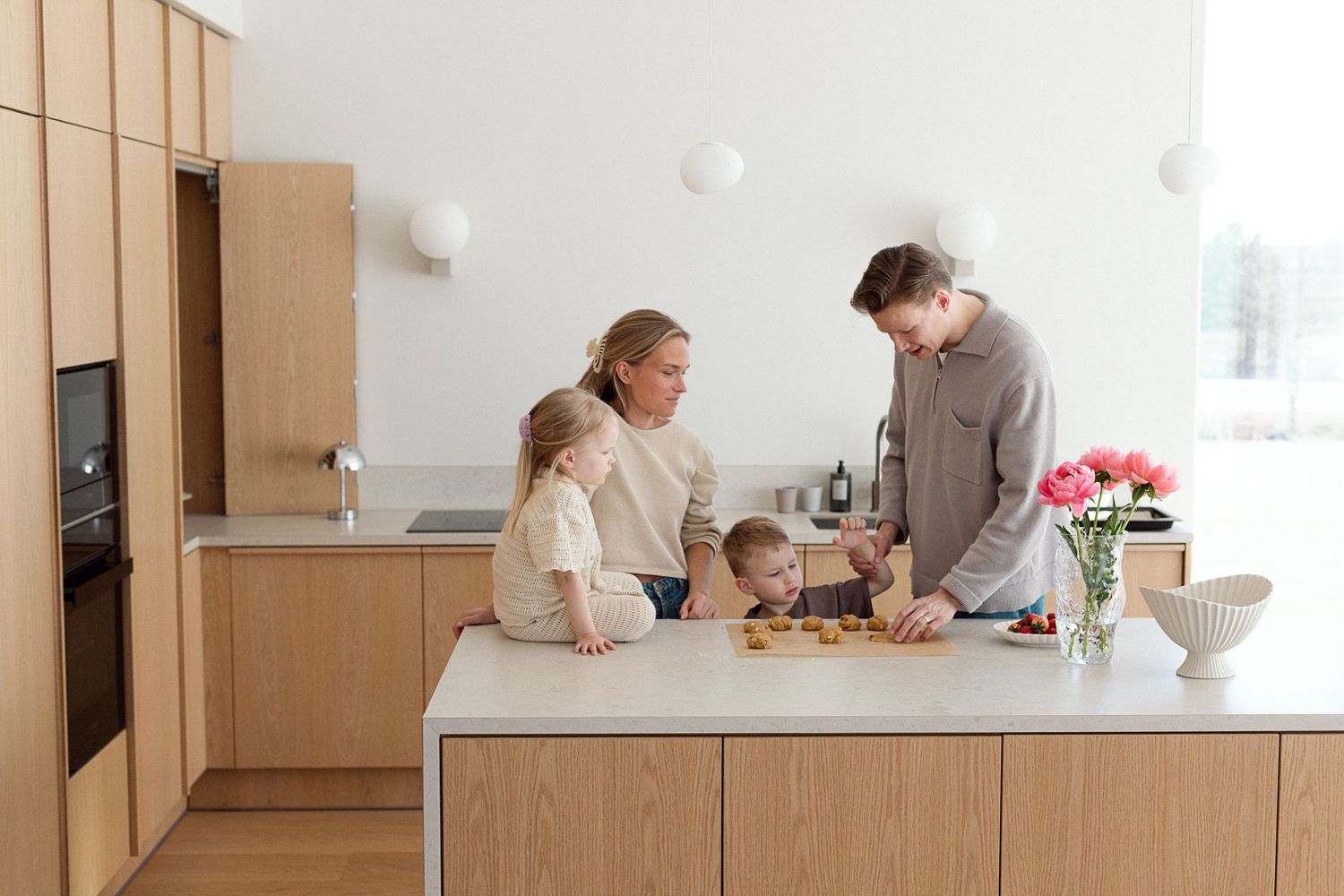 Family cooking together in a modern Scandinavian kitchen with wooden cabinets and white countertops.
