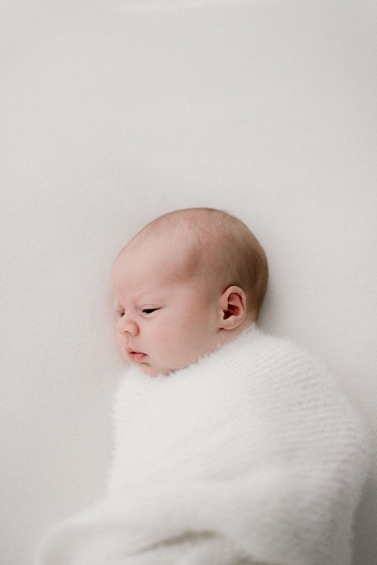 A peaceful newborn baby swaddled in a soft white knit wrap, resting against a white background.