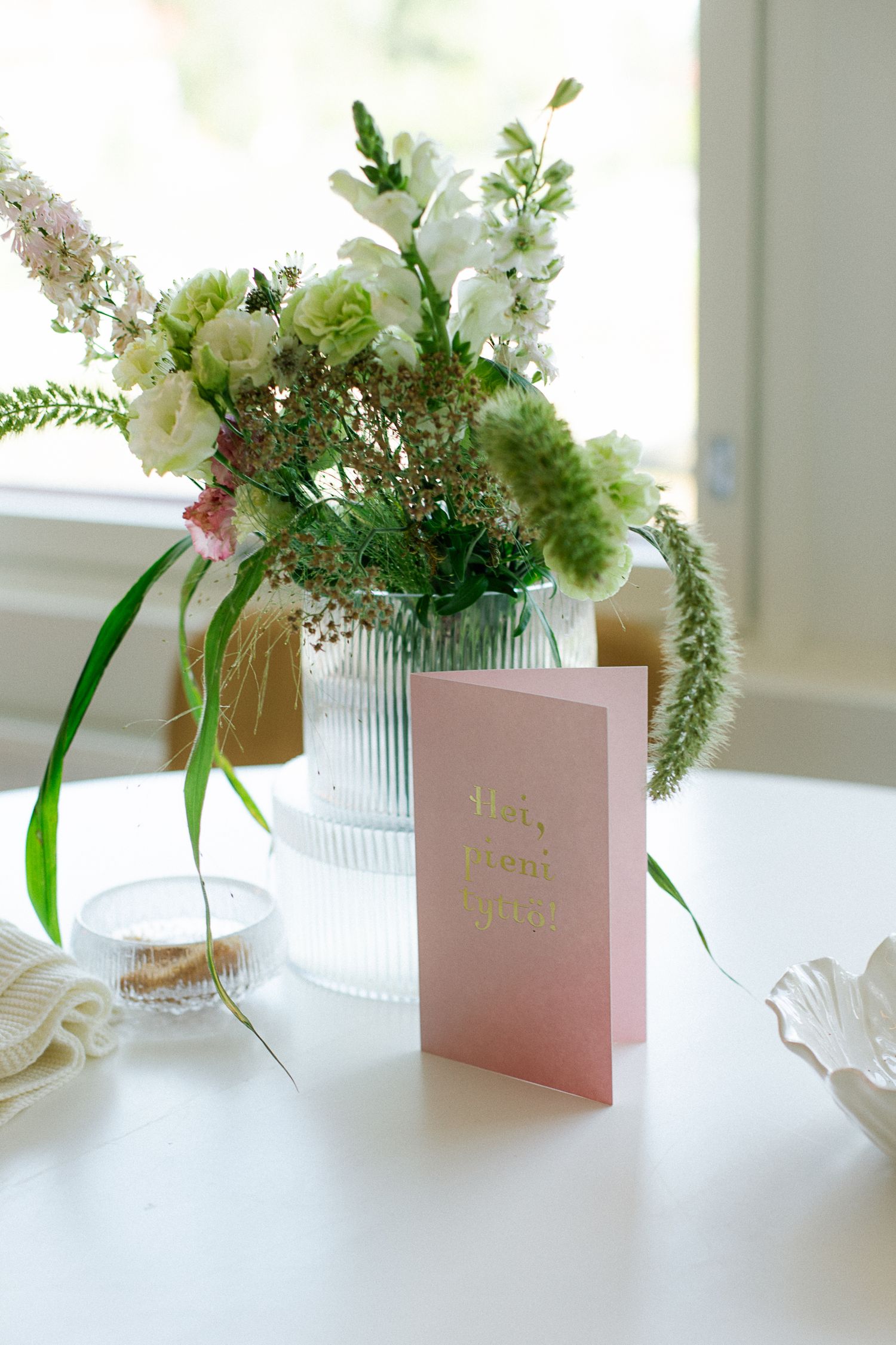 A glass ribbed vase with white and green flowers on a white table beside a pink greeting card.