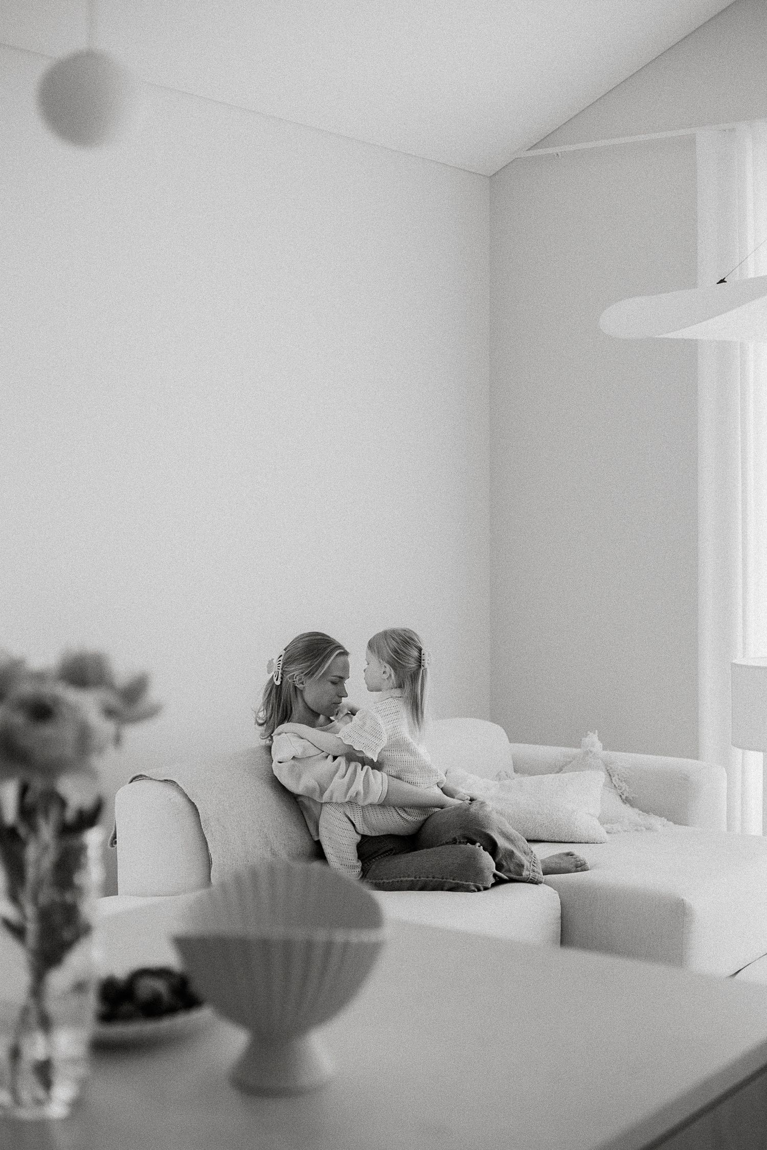Black and white photo of two young girls sitting together on a white sofa in a bright minimalist living room.