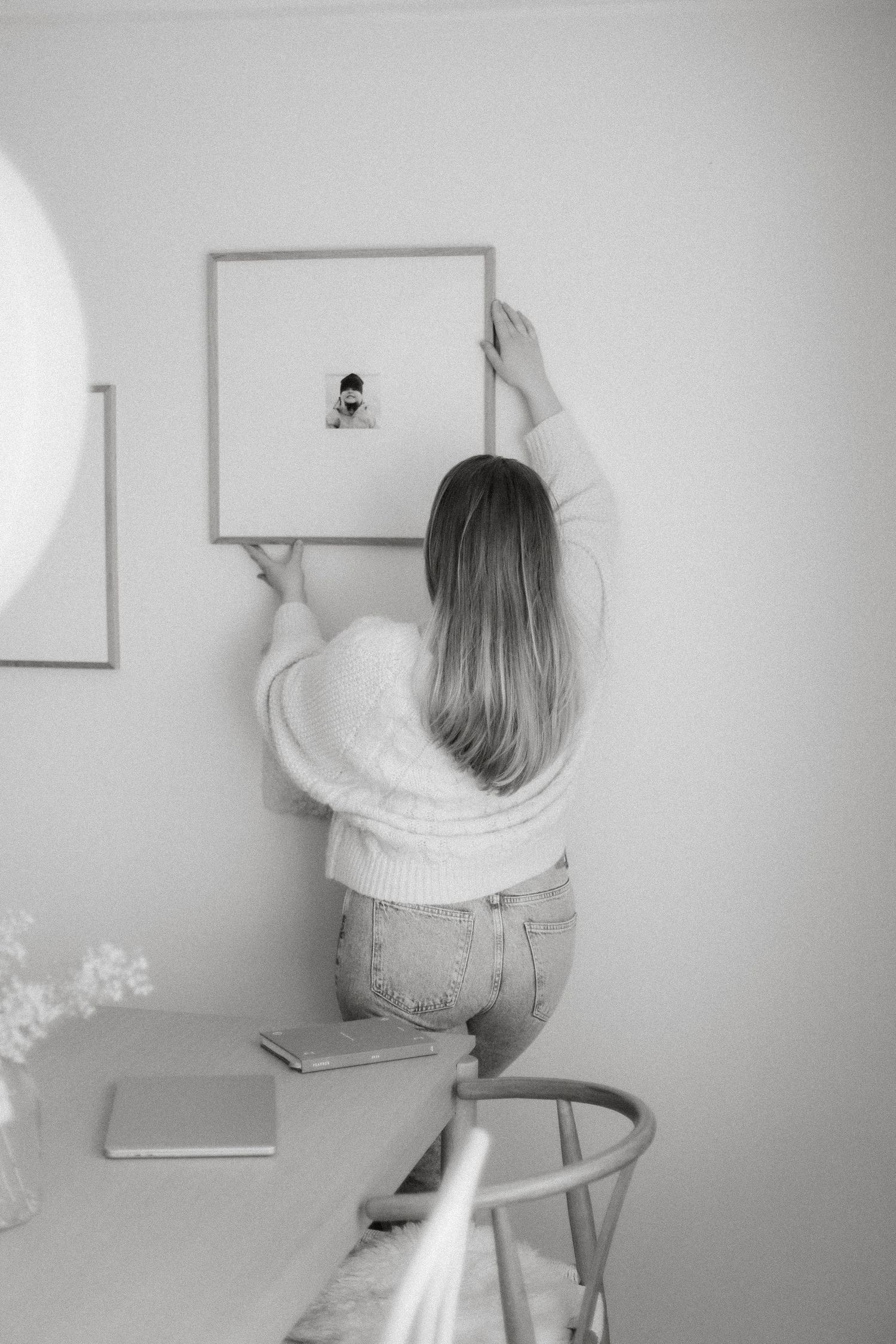 Black and white photo of a woman hanging a framed picture on a white wall in a minimalist room.