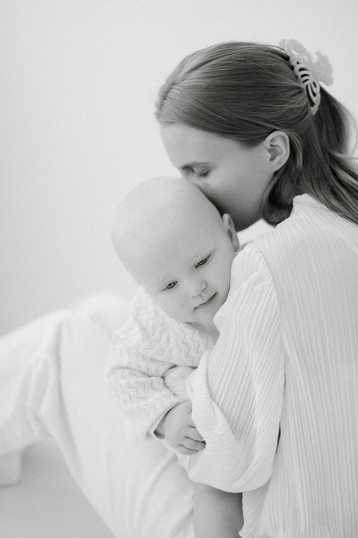 Black and white photo of a mother tenderly kissing her smiling baby on the head while gently holding them.