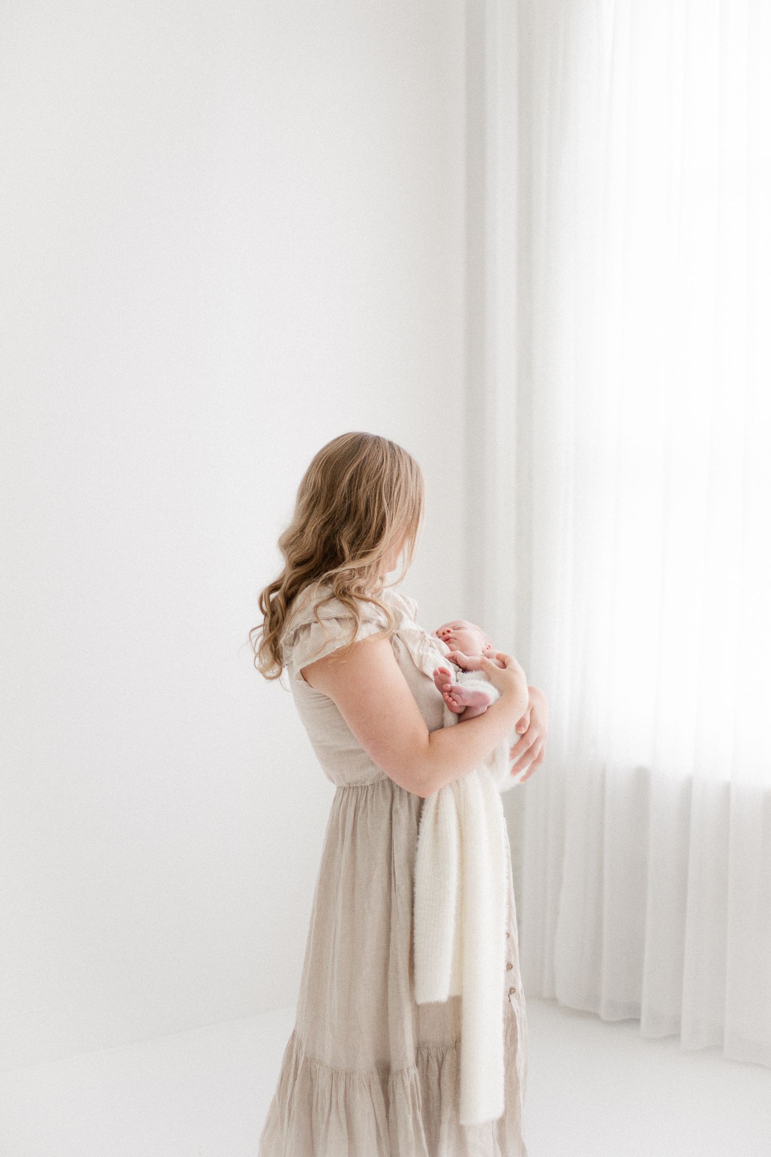 A mother in a beige dress holds her newborn baby wrapped in white near a bright window.