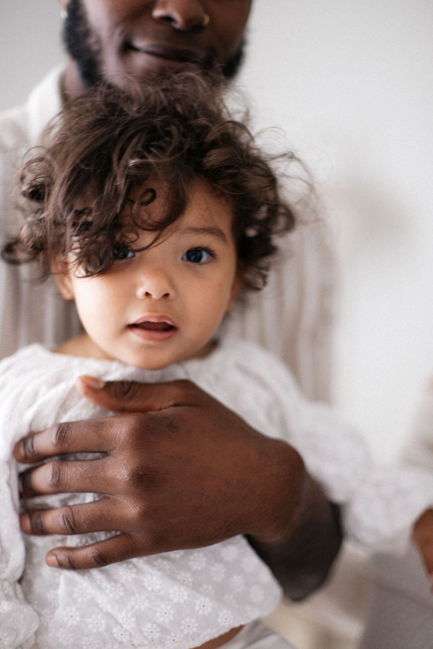 A man with dark skin tenderly holds a curly-haired toddler in a white dress against a light background.