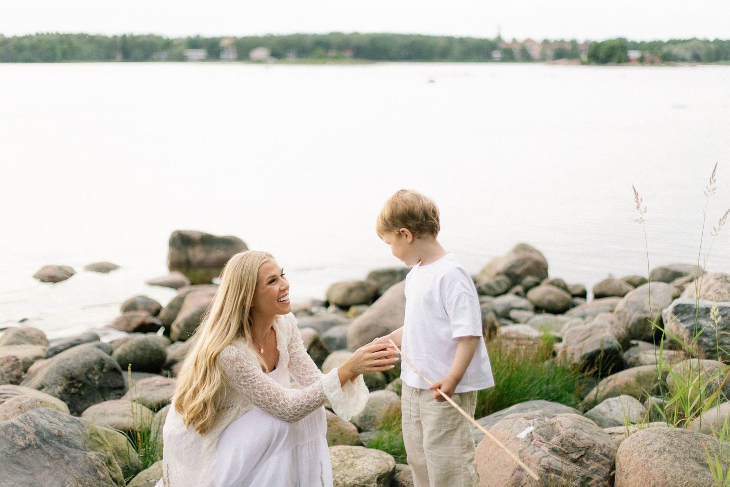 A woman in a white dress talks with a boy on a rocky shoreline near calm water.