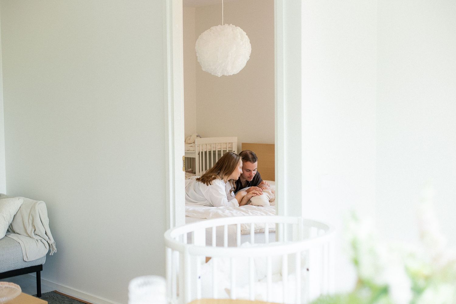 Parents lying on the floor bonding with their newborn baby in a bright white nursery room with a white crib.