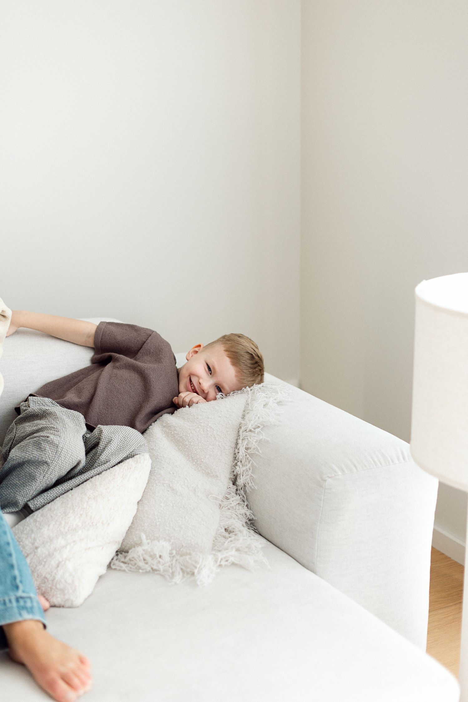 A happy young child laughing while lying on a white sofa with pillows in a bright, minimalist living room.