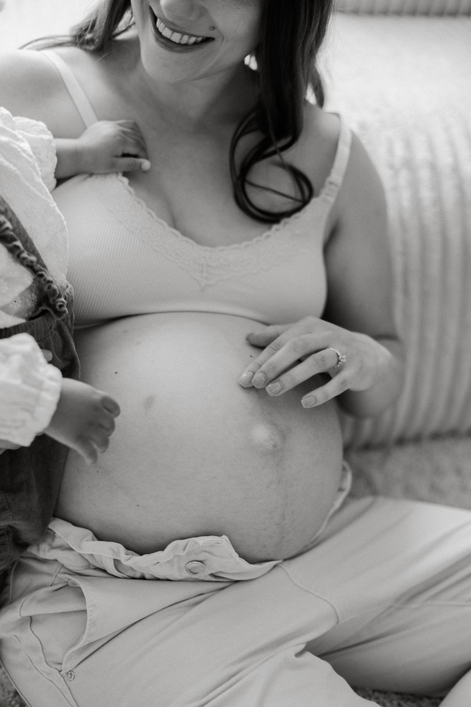 Black and white maternity photo of smiling pregnant woman in bra cradling bare baby bump while seated.