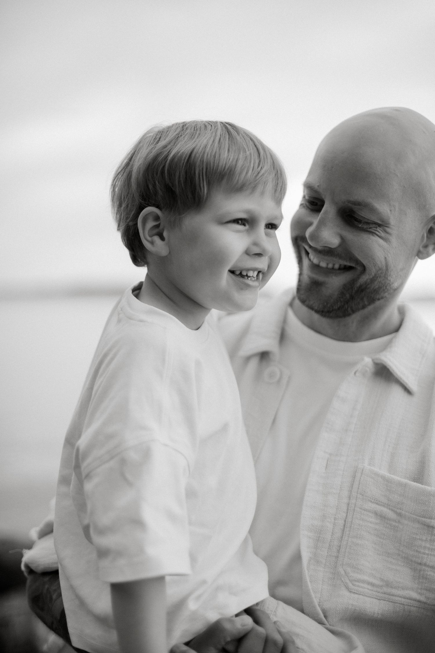 Black and white photo of a smiling bald father holding his laughing young son outdoors near water.