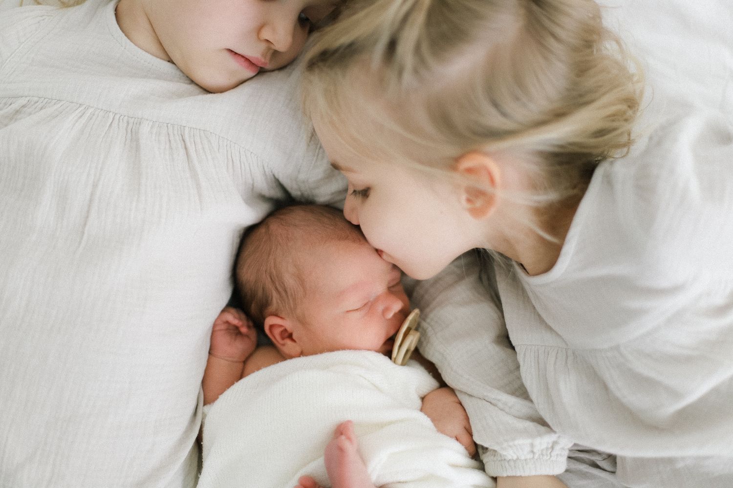 Two young girls in white dresses tenderly cuddle a sleeping newborn baby wrapped in a white blanket.