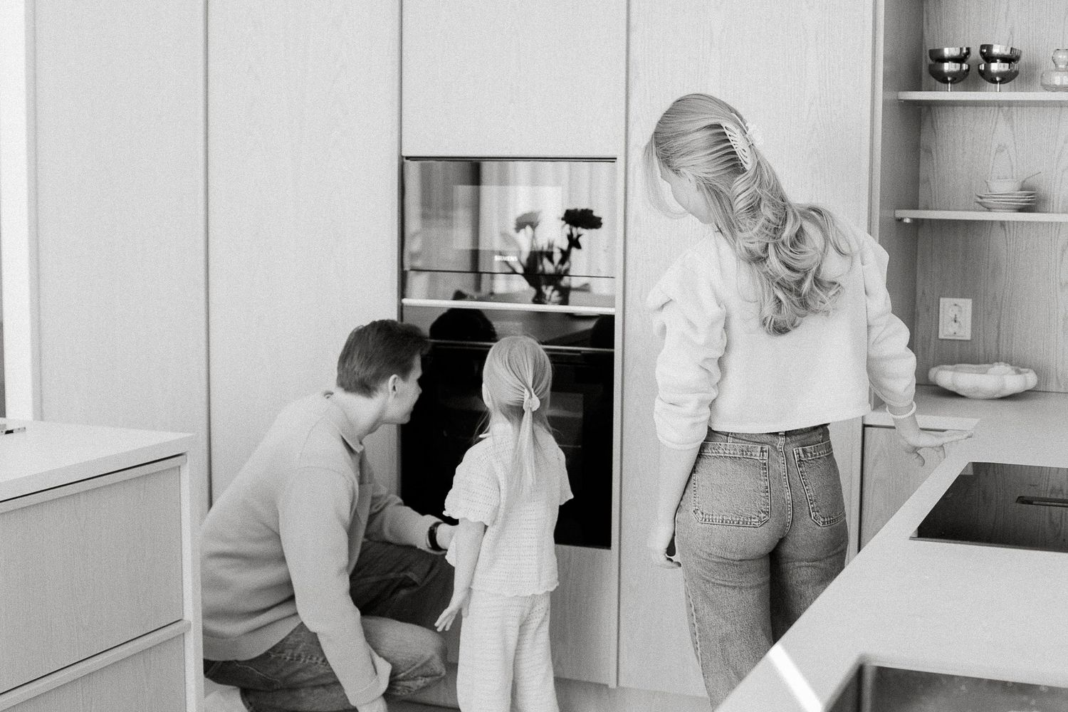 Black and white photo of a family with a young child looking into an oven together in a modern kitchen.