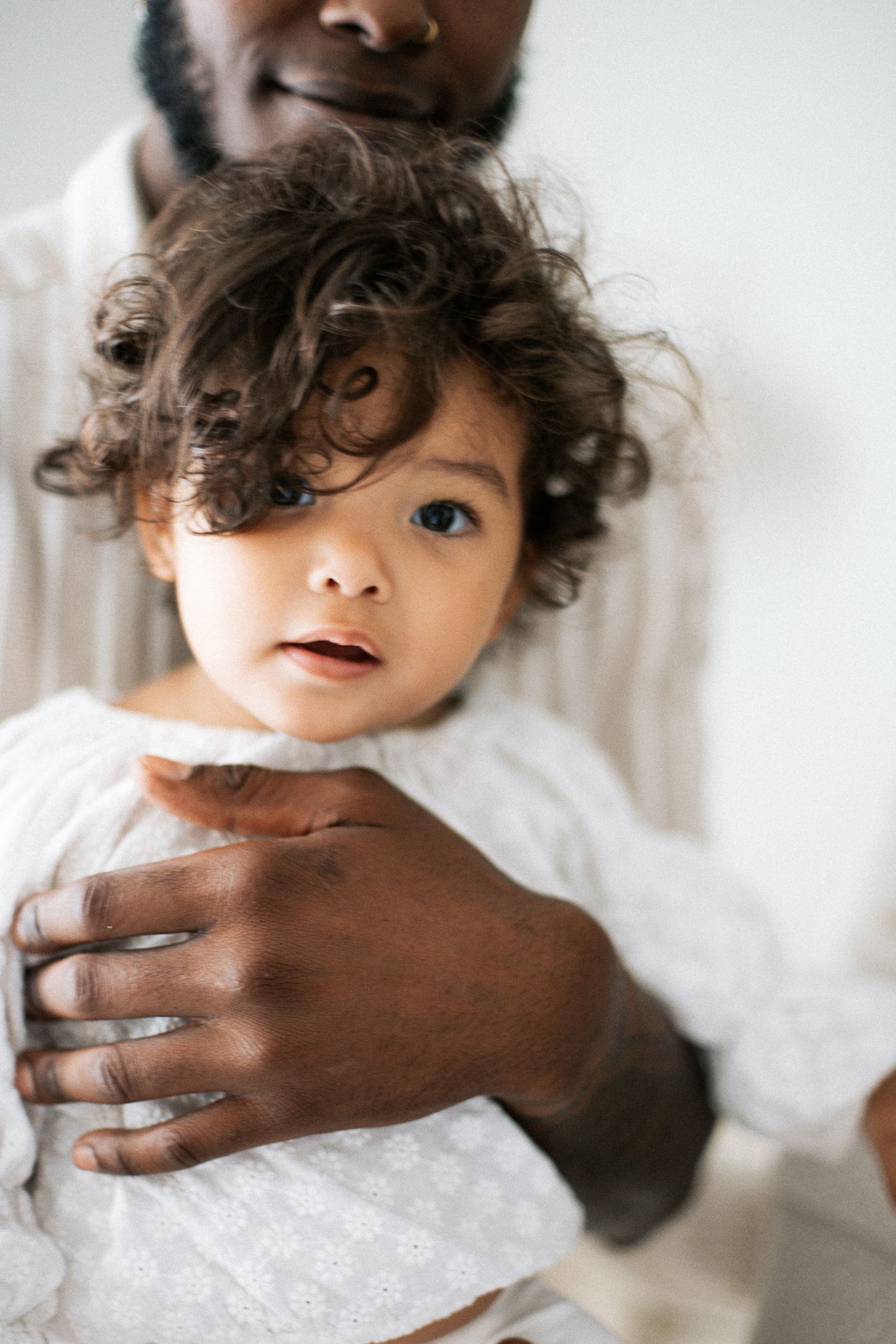A Black father tenderly holds his curly-haired toddler dressed in white, sharing a gentle moment together.