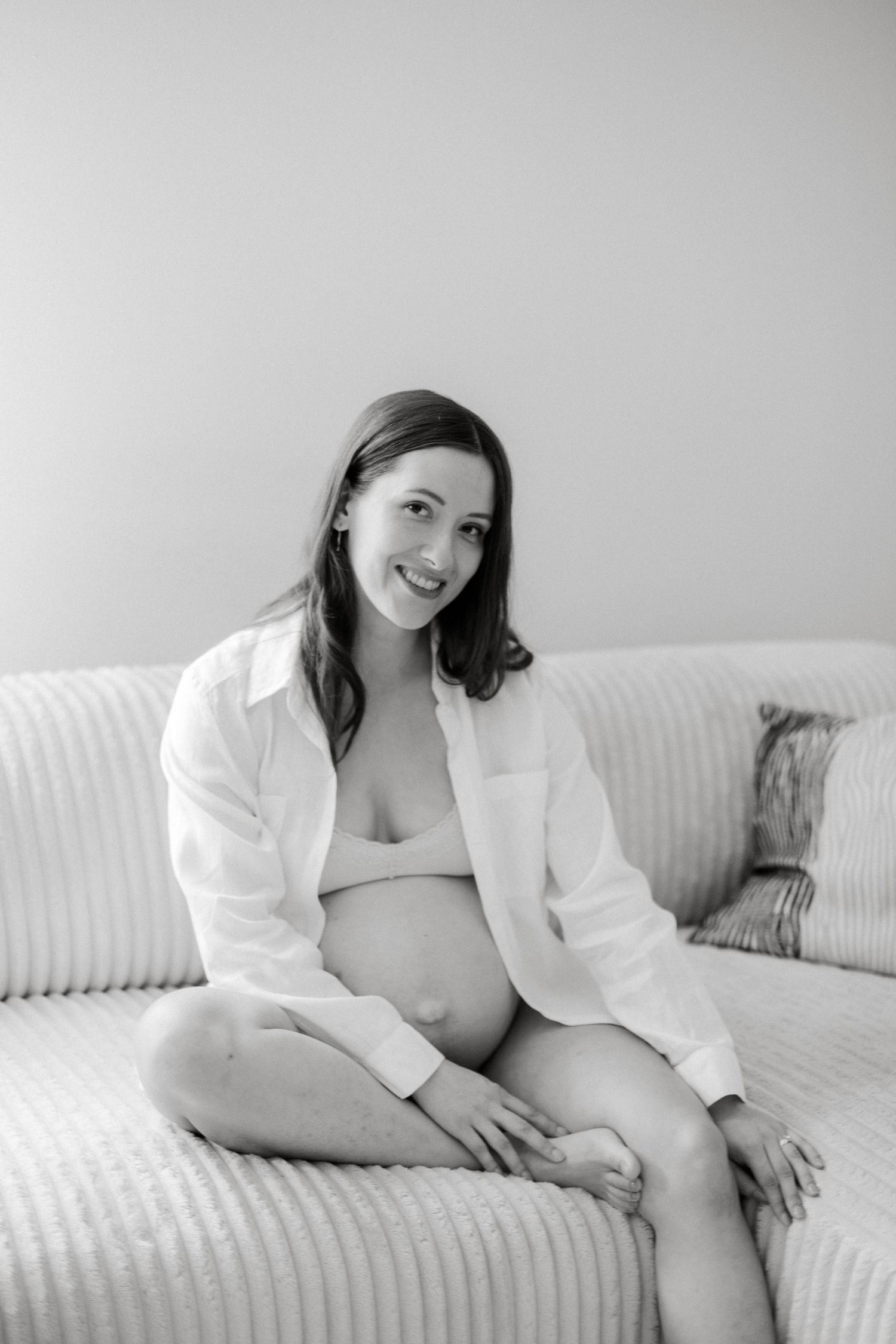Black and white photo of a smiling pregnant woman sitting cross-legged on a sofa, wearing an open white shirt.