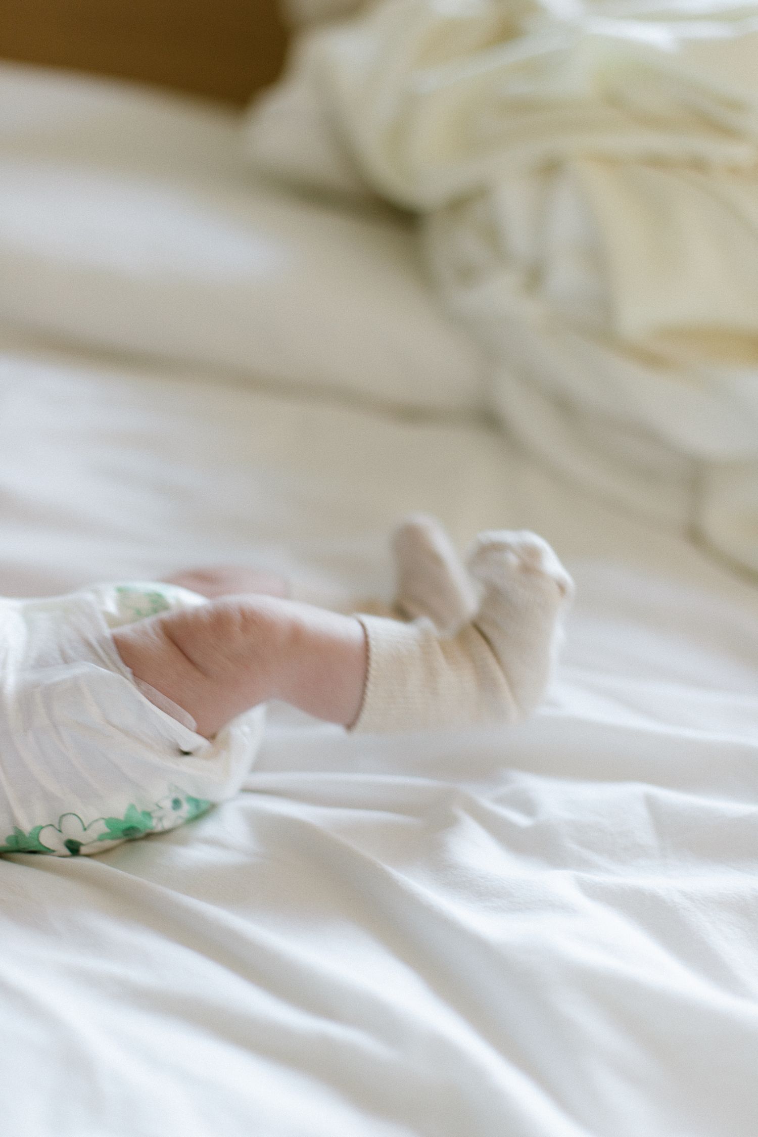 Close-up of baby feet in white socks lying on a white bed, wearing a floral onesie.