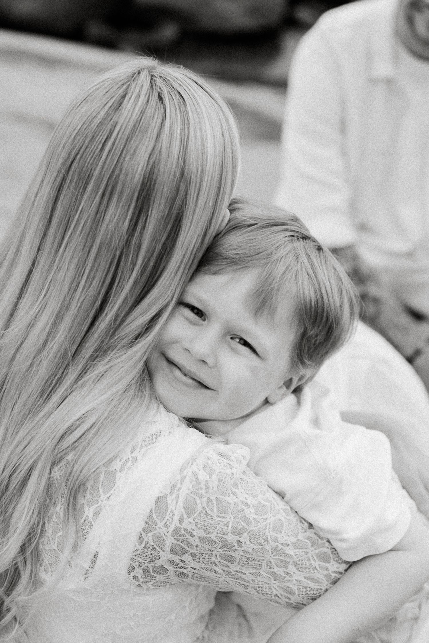 Black and white photo of a young child smiling while being hugged by an older girl with long blonde hair.