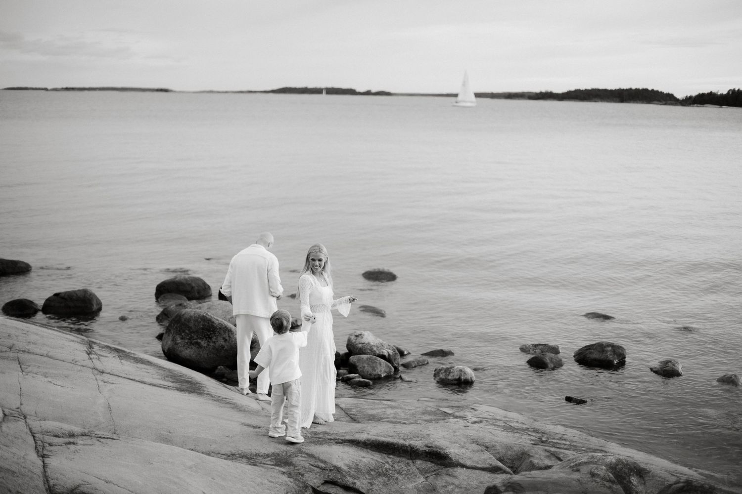 Black and white photo of a family in white clothing standing on rocky shoreline gazing at calm sea with sailboat.