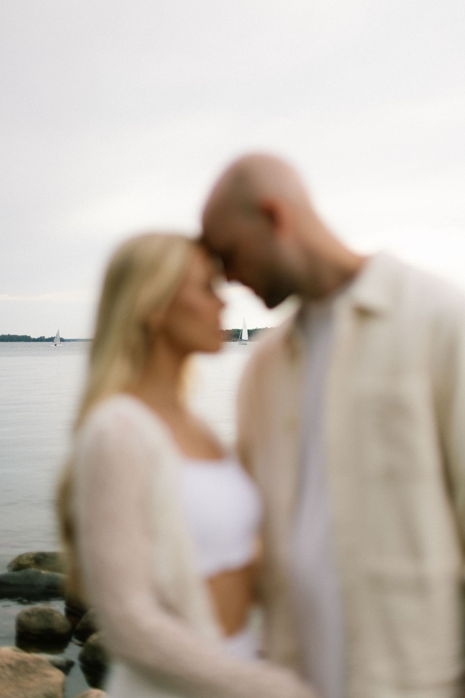 A blurred romantic couple touching foreheads by a serene waterfront with rocks and a sailboat in the background.