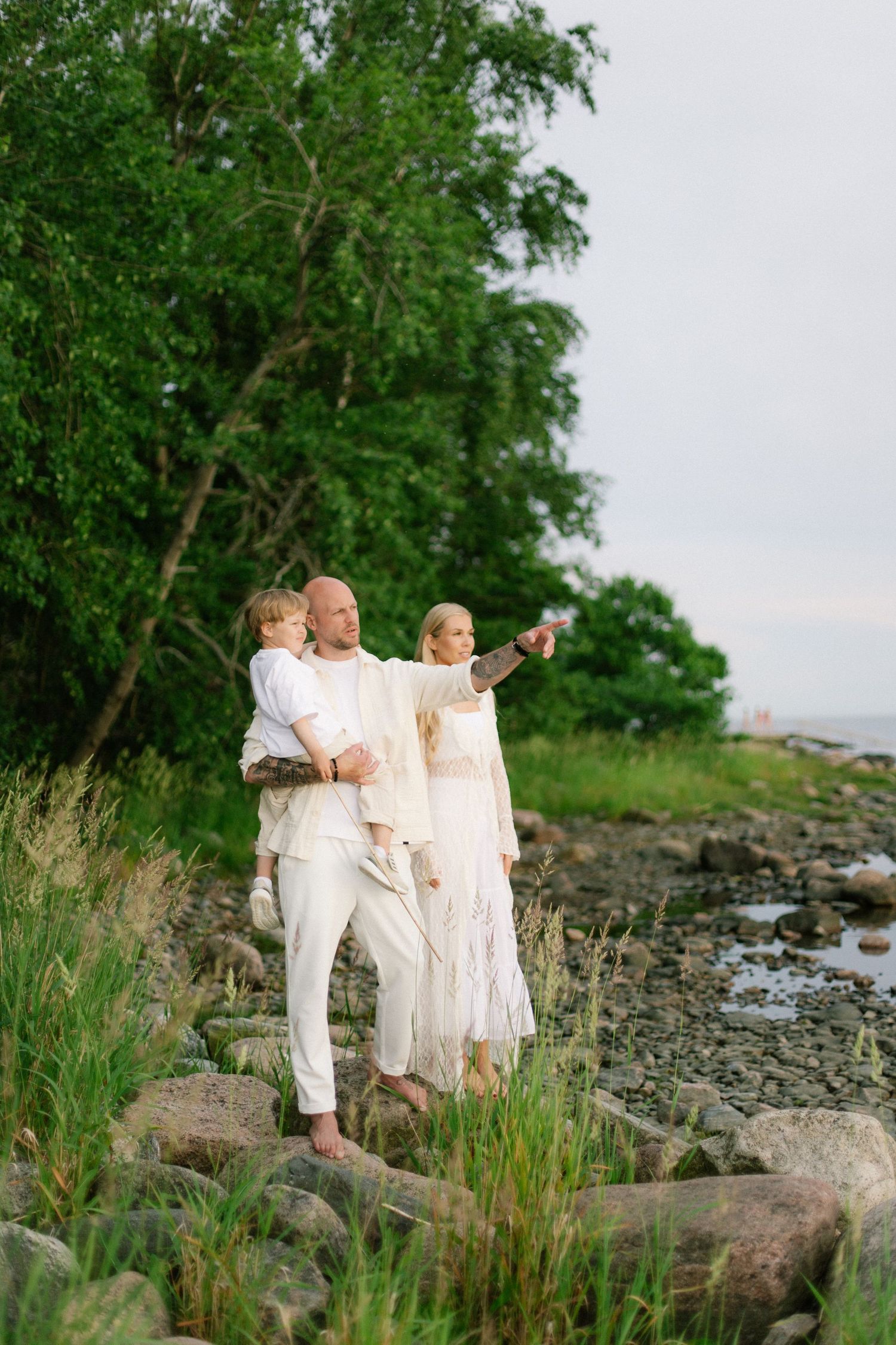 Family in white outfits standing on rocky shore, man holding child while woman points toward horizon near green trees.
