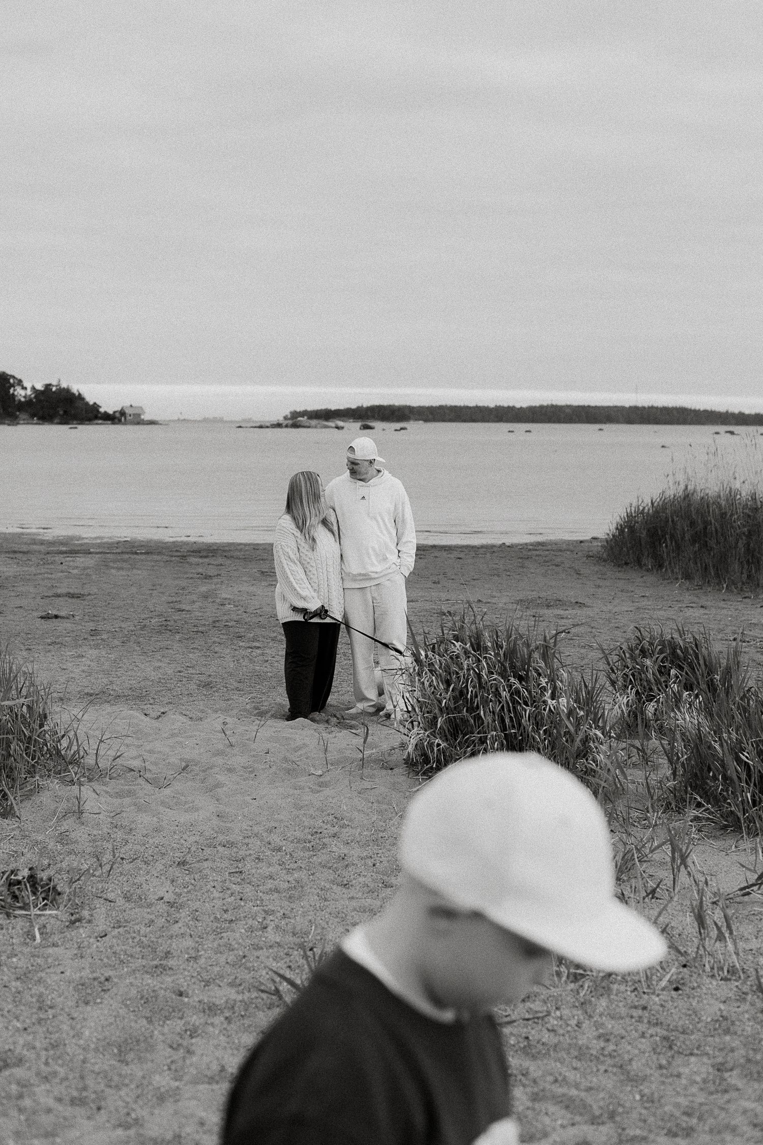 Black and white photo of a couple standing on a sandy beach shore, with a person in a cap in the foreground.