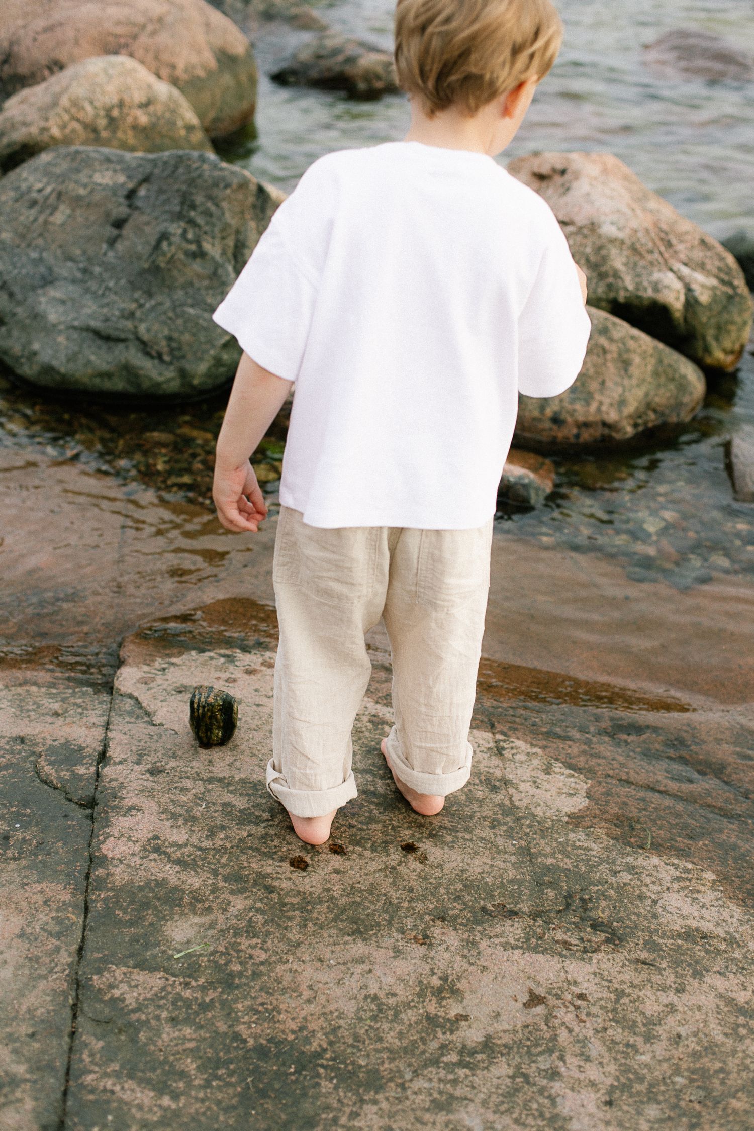Young barefoot child in white t-shirt and linen pants stands on rocky beach exploring the shoreline.