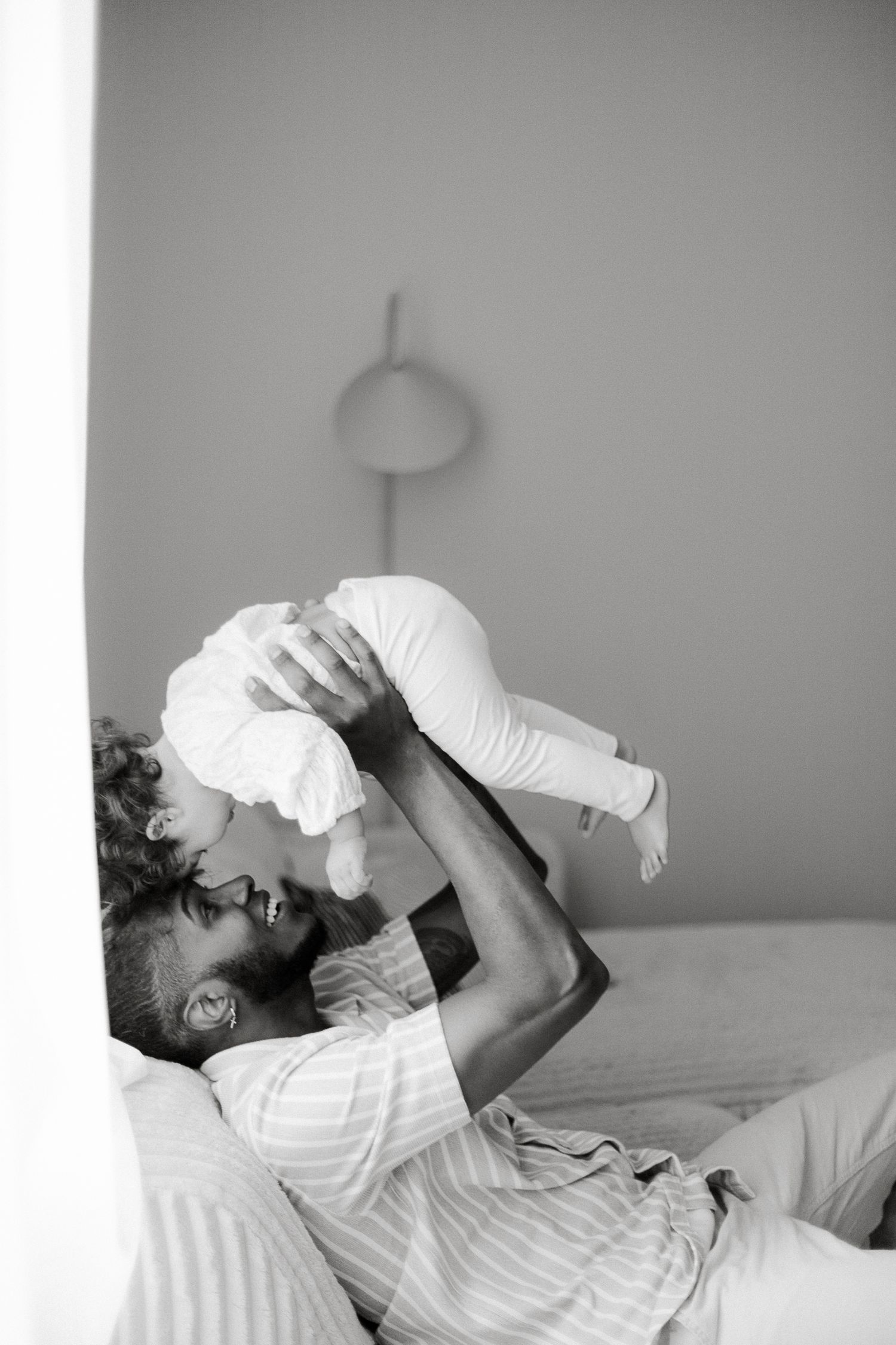 Black and white photo of a parent lying on bed, joyfully lifting their baby up in the air.
