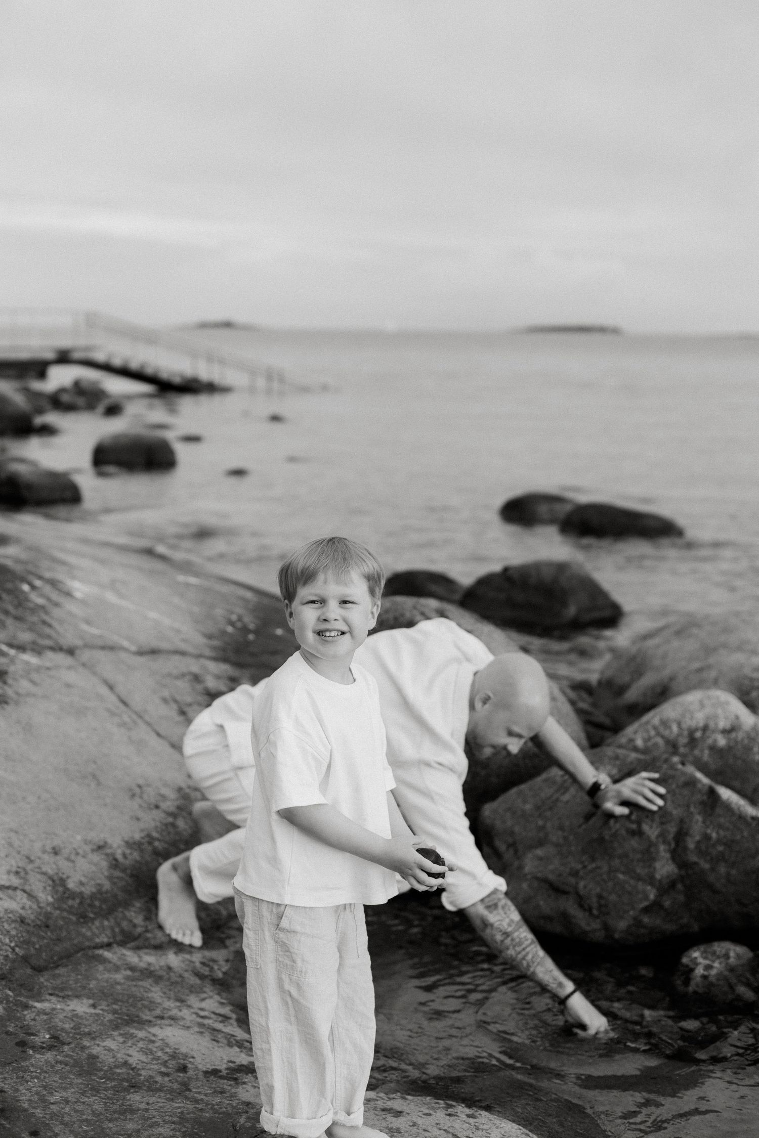 Black and white photo of a smiling child in white clothes on rocky shoreline with another child climbing rocks nearby.