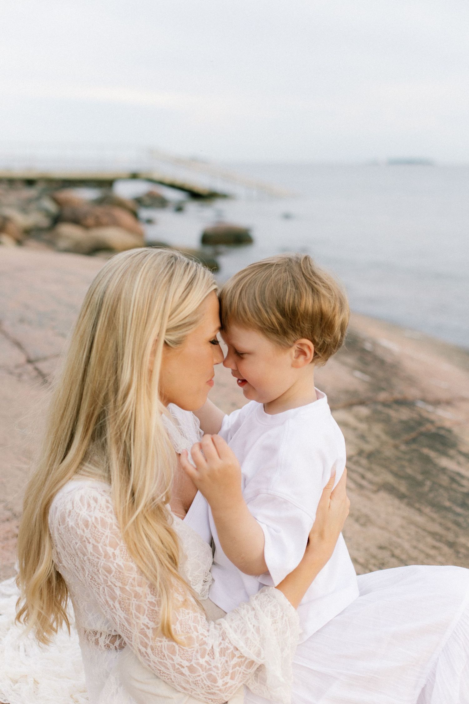 A blonde mother in white lace holds her young son close, touching foreheads tenderly on a rocky seaside shore.