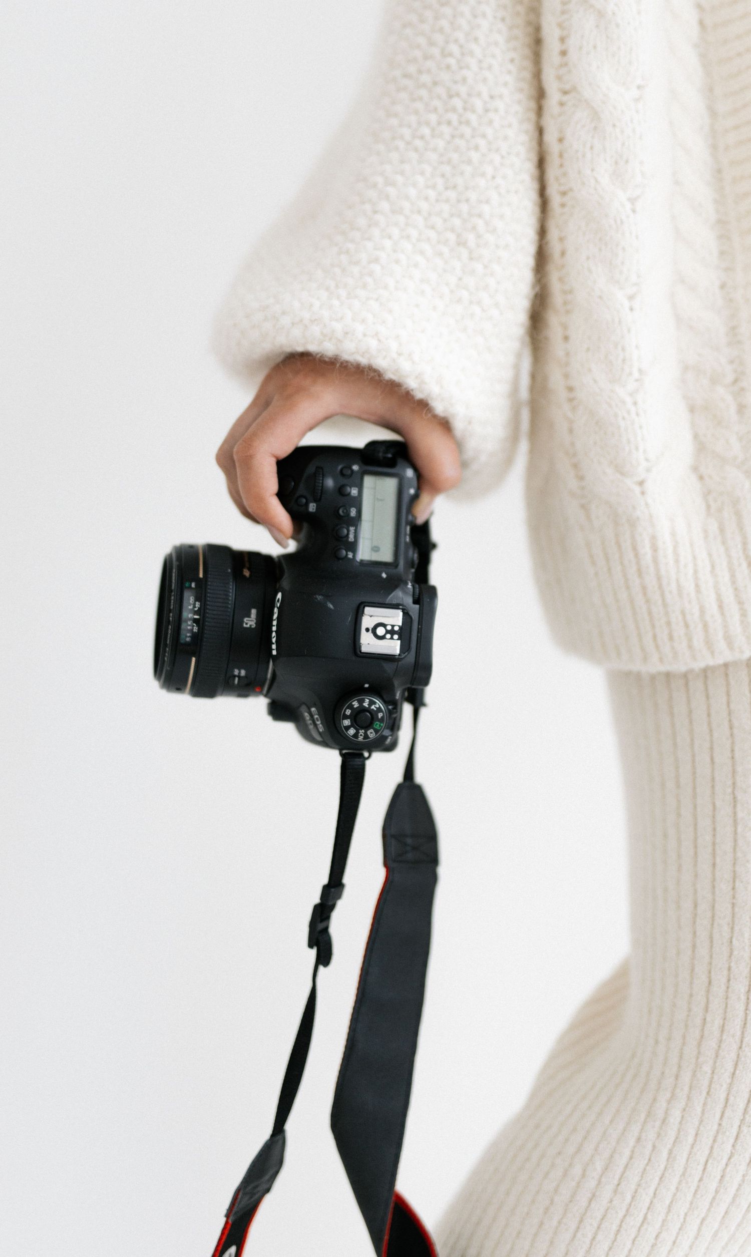 Person in cream cable-knit sweater holding a Canon DSLR camera with strap against white background.