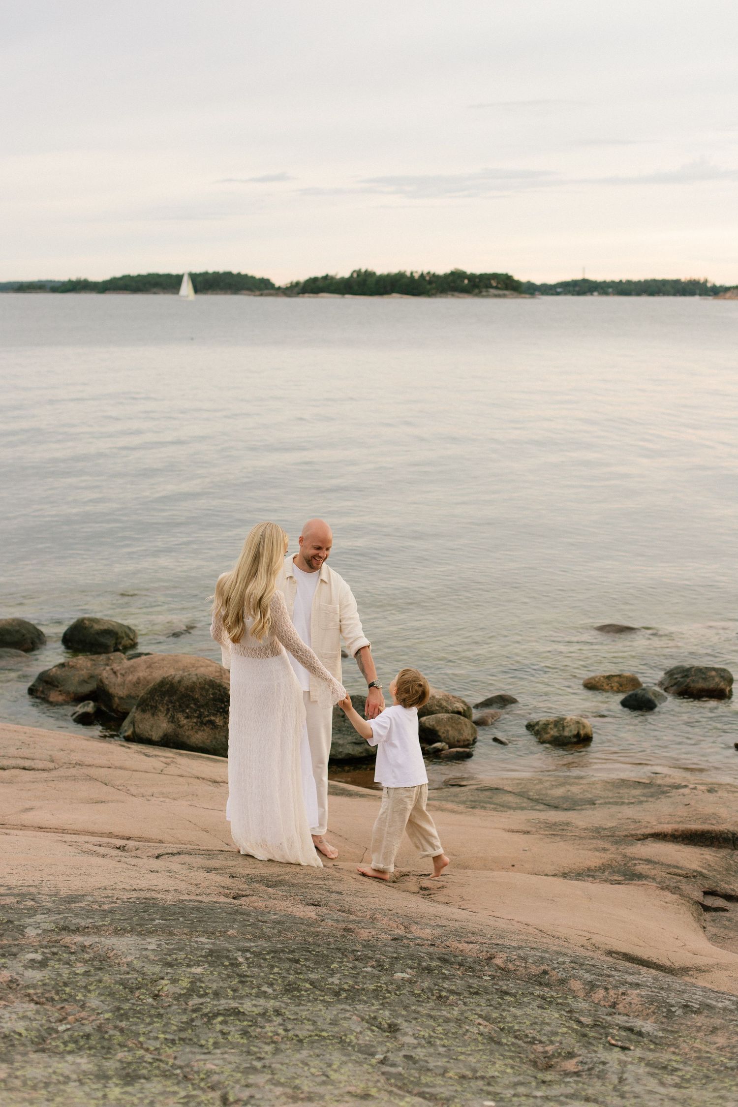 Family in white outfits holding hands on rocky shoreline with calm sea and island in background.