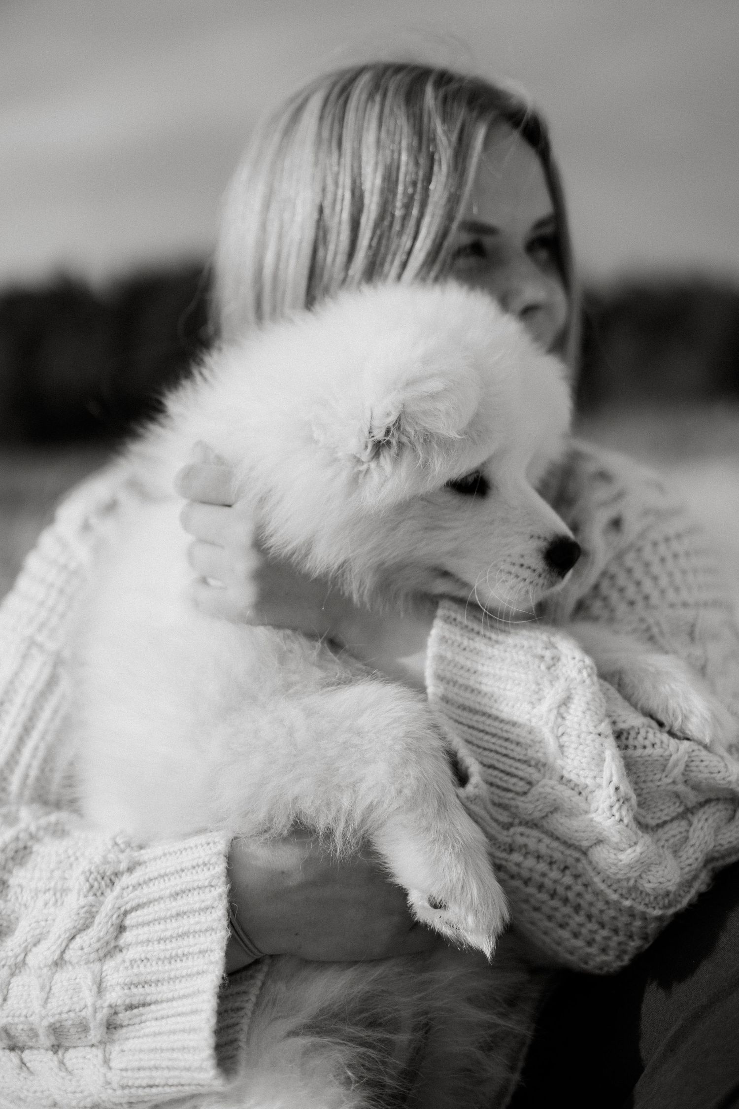 Black and white photo of a woman in a knit sweater tenderly holding a fluffy white Samoyed puppy outdoors.