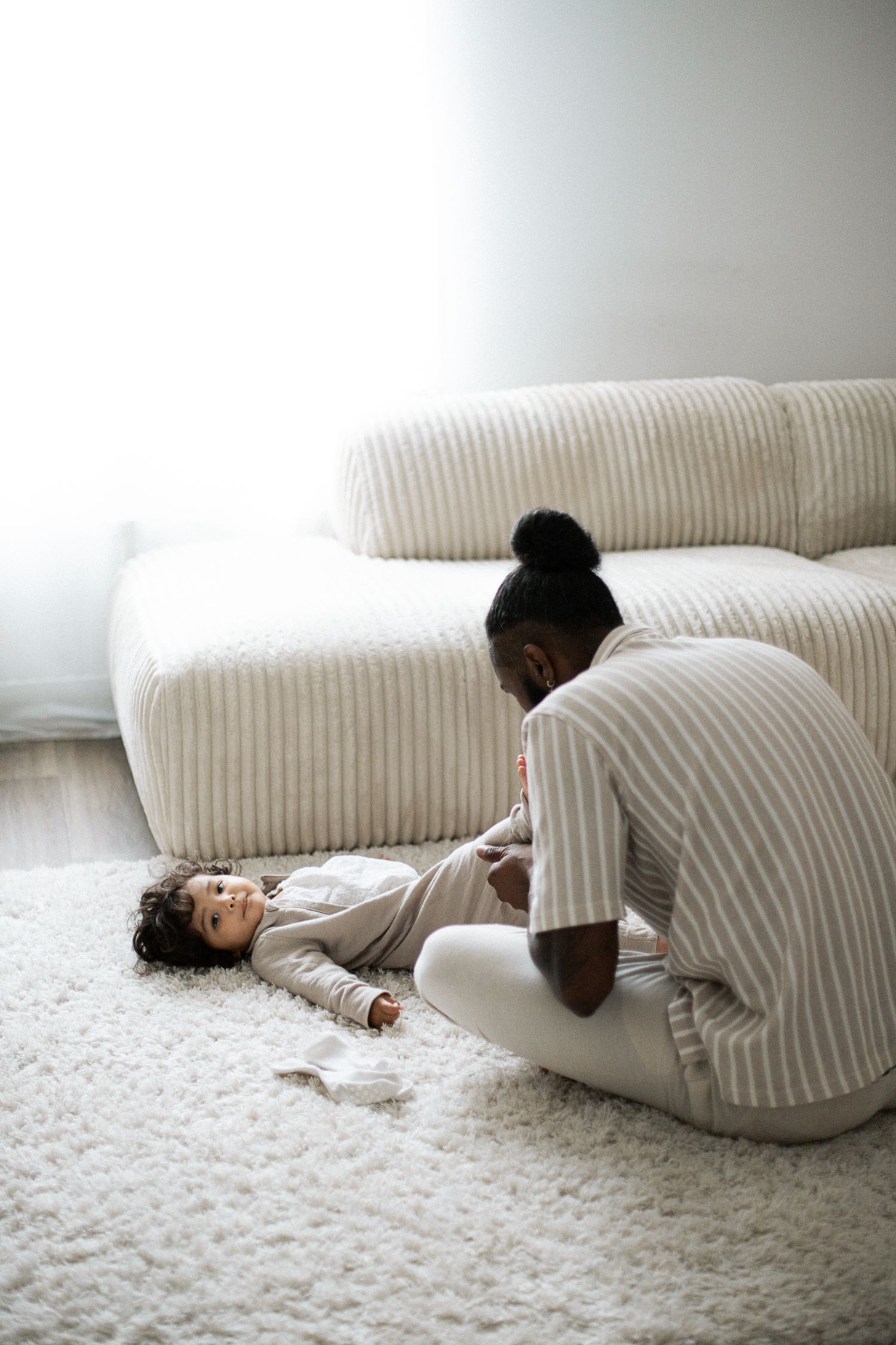 A parent in striped pajamas plays with a baby on a fluffy white rug beside a modern cream sofa.