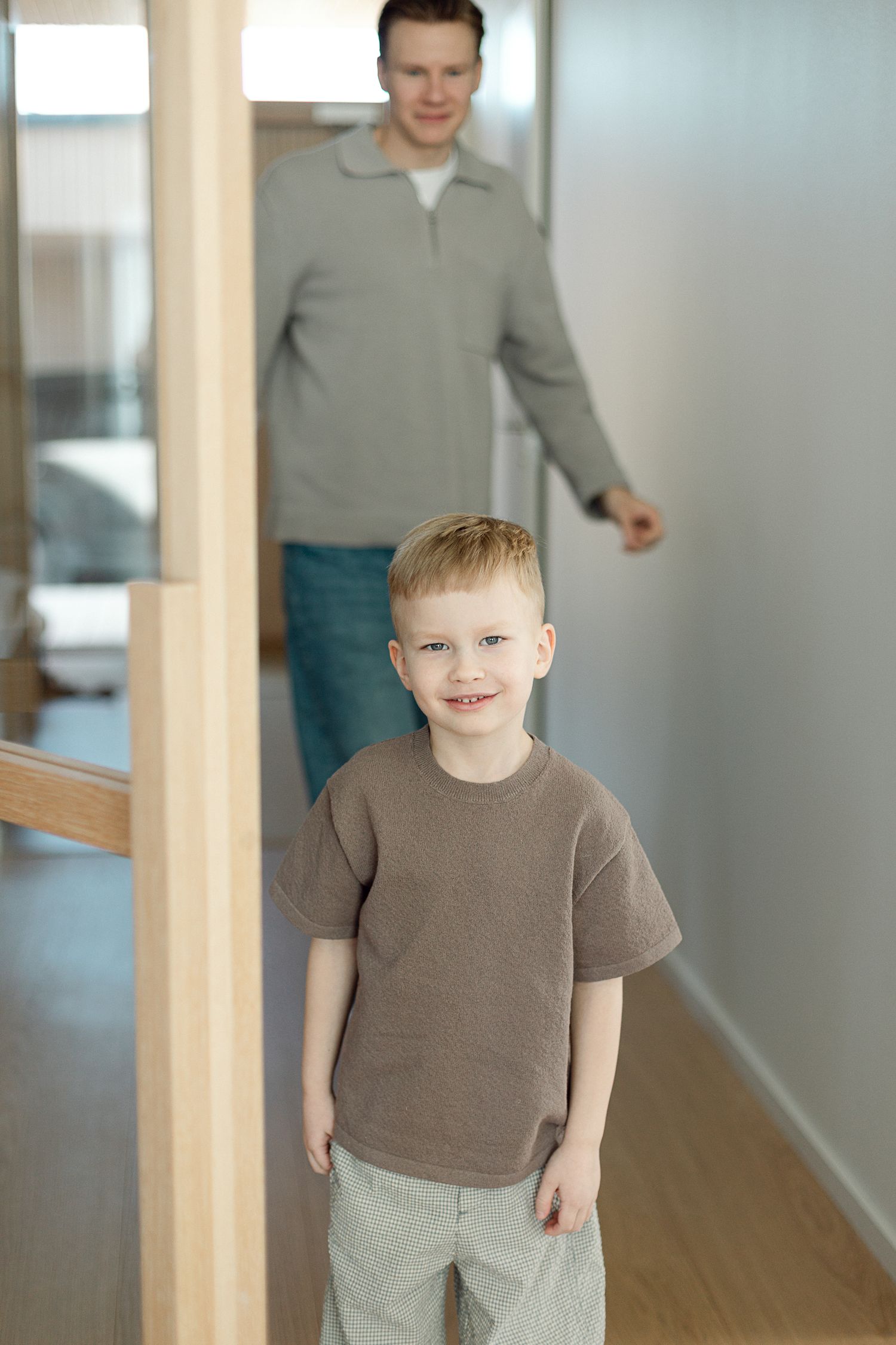 A smiling young boy in a brown t-shirt stands in a hallway with an adult man visible in the background.