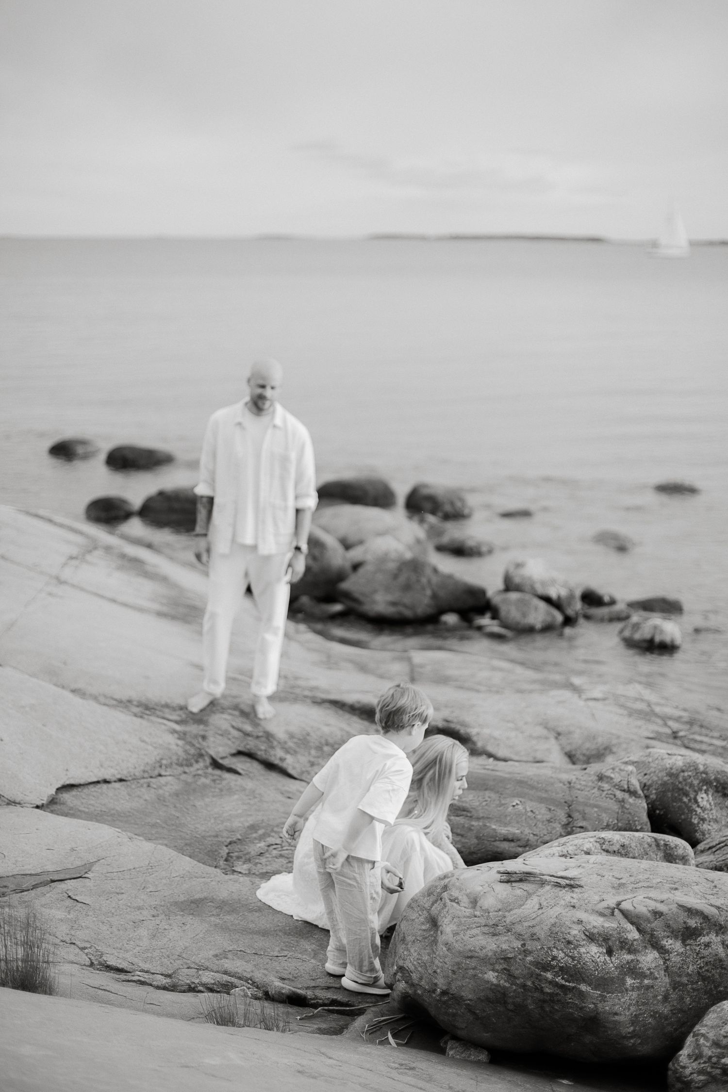 Black and white photo of a family on rocky beach, man standing by water while two children crouch on rocks.