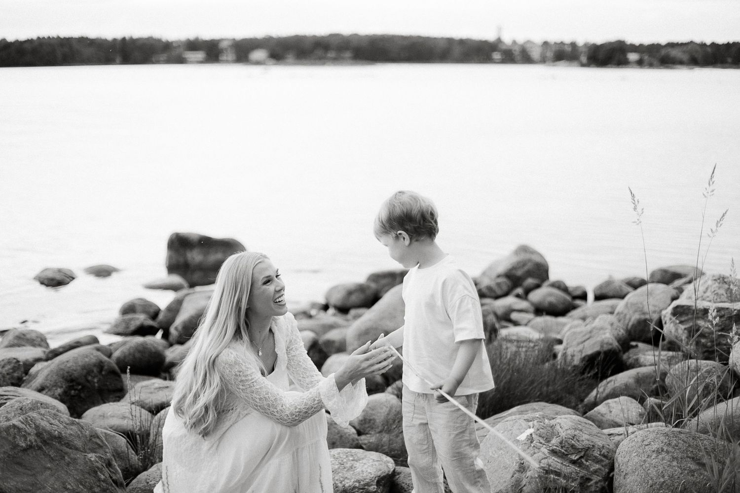 Black and white photo of a woman and child on rocky shoreline near calm water.