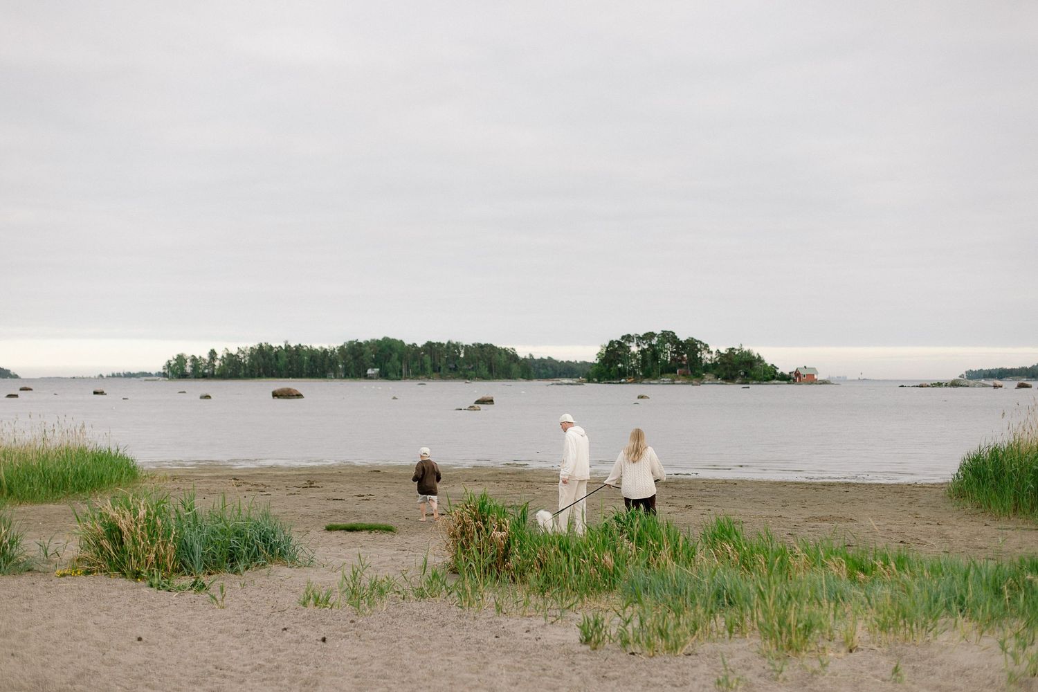 Two people and a child walk on a sandy beach toward calm waters with small islands in the distance.