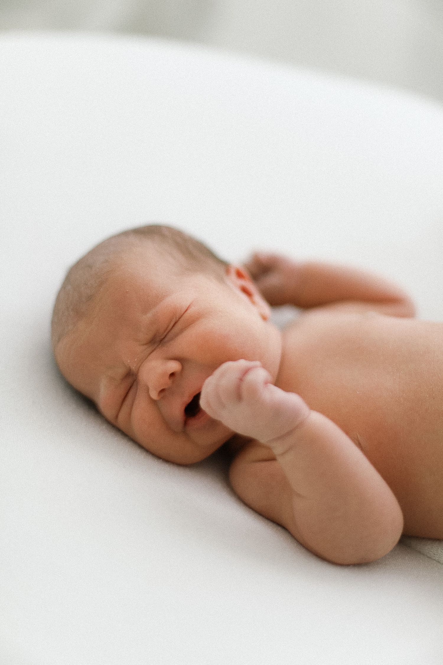 Newborn baby lying on white surface with eyes closed and fist near face, peaceful sleeping pose.