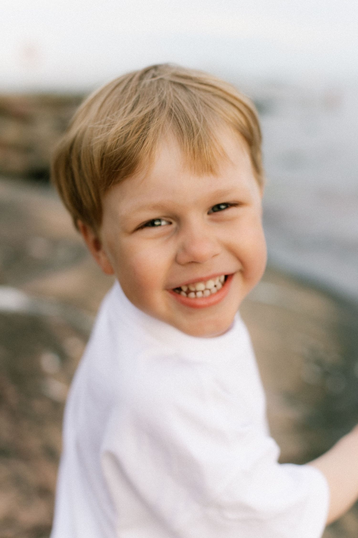 Smiling young boy with blonde hair wearing a white t-shirt near a rocky shoreline.