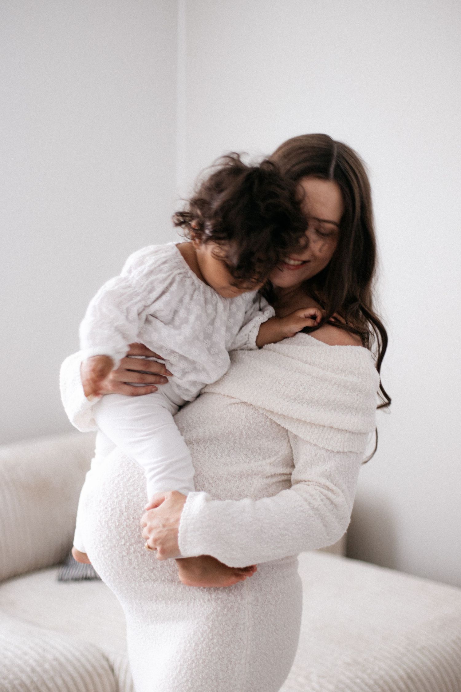 A smiling pregnant woman in a white dress lovingly holds a curly-haired toddler in a bright, minimalist room.
