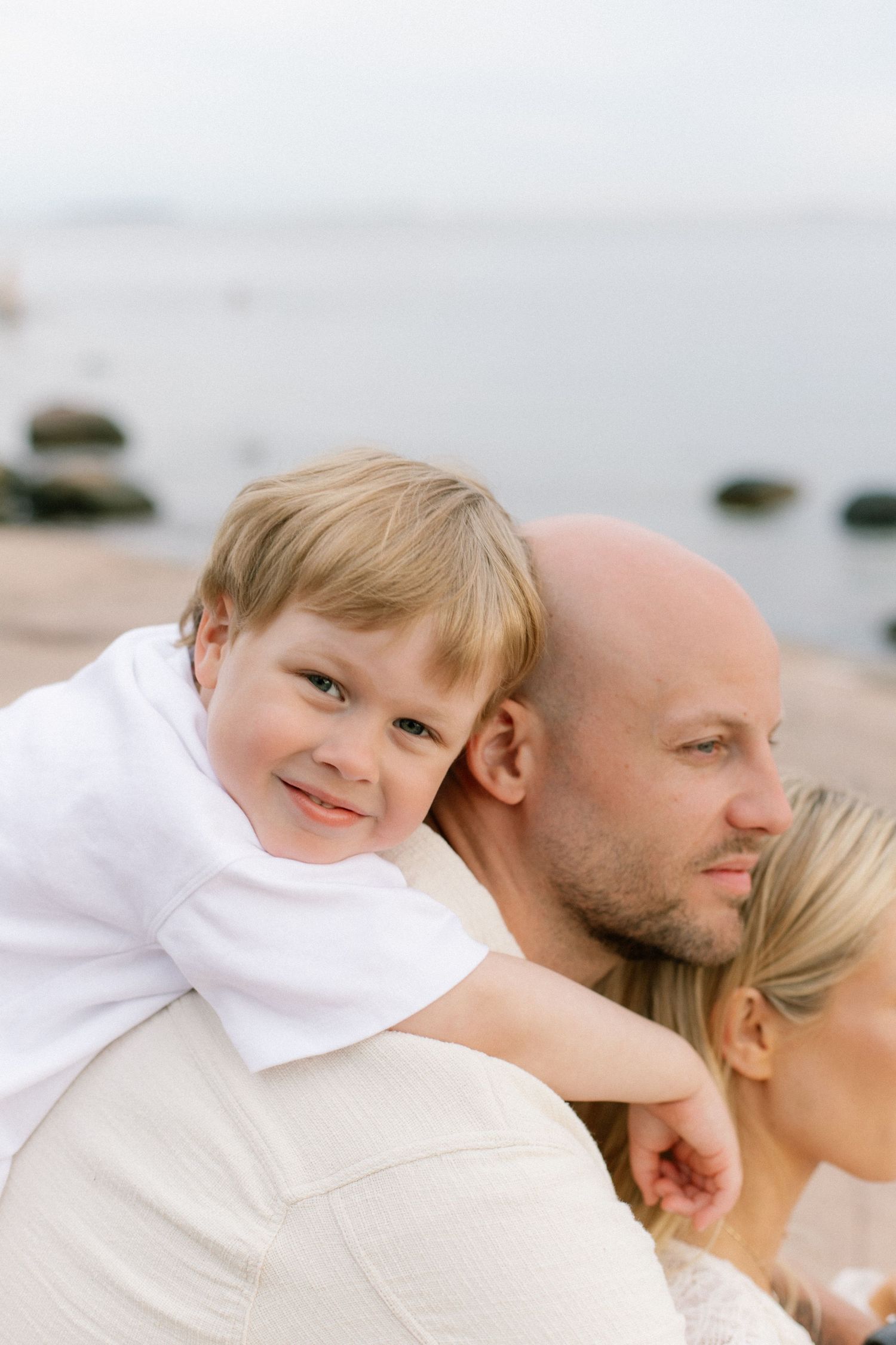 A young boy smiles on his father's shoulders at a beach, with a woman beside them on a cloudy day.
