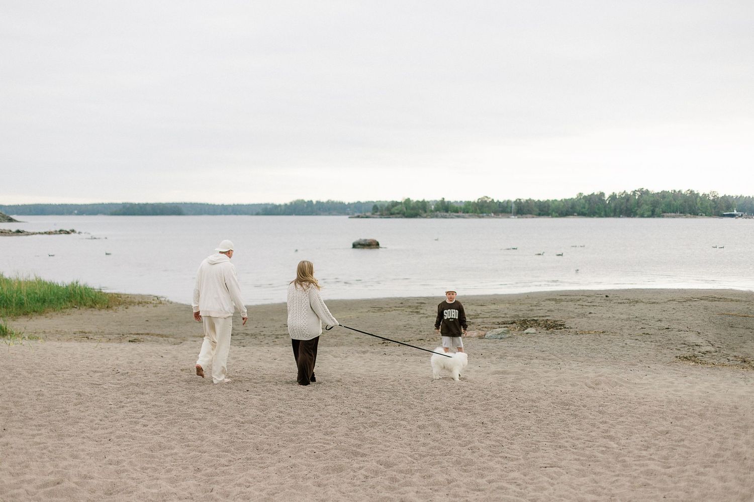 Family walking a white dog on a sandy beach with calm water and green trees in the background on an overcast day.