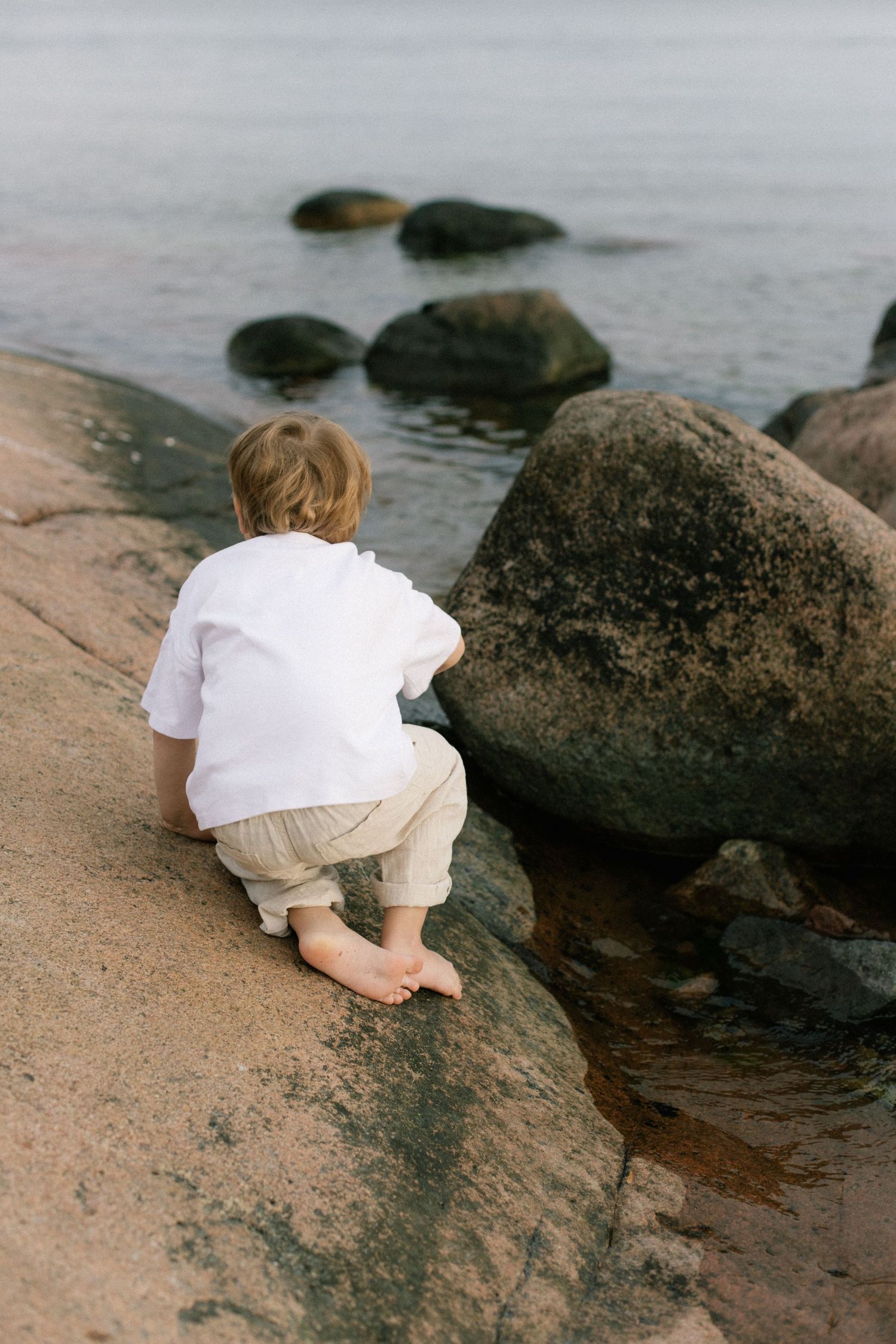 Young barefoot child crouching on rocky shoreline, exploring stones by calm water's edge.