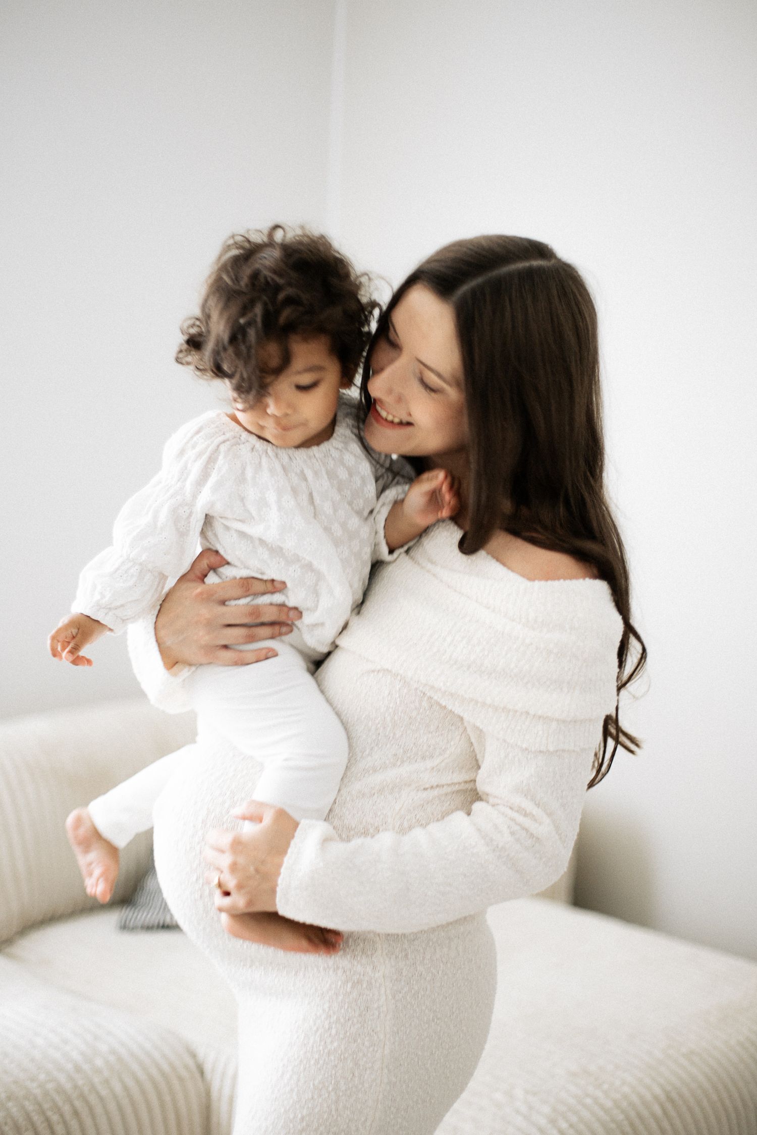 A smiling mother in white dress lovingly holds her curly-haired toddler in a bright, minimalist room.