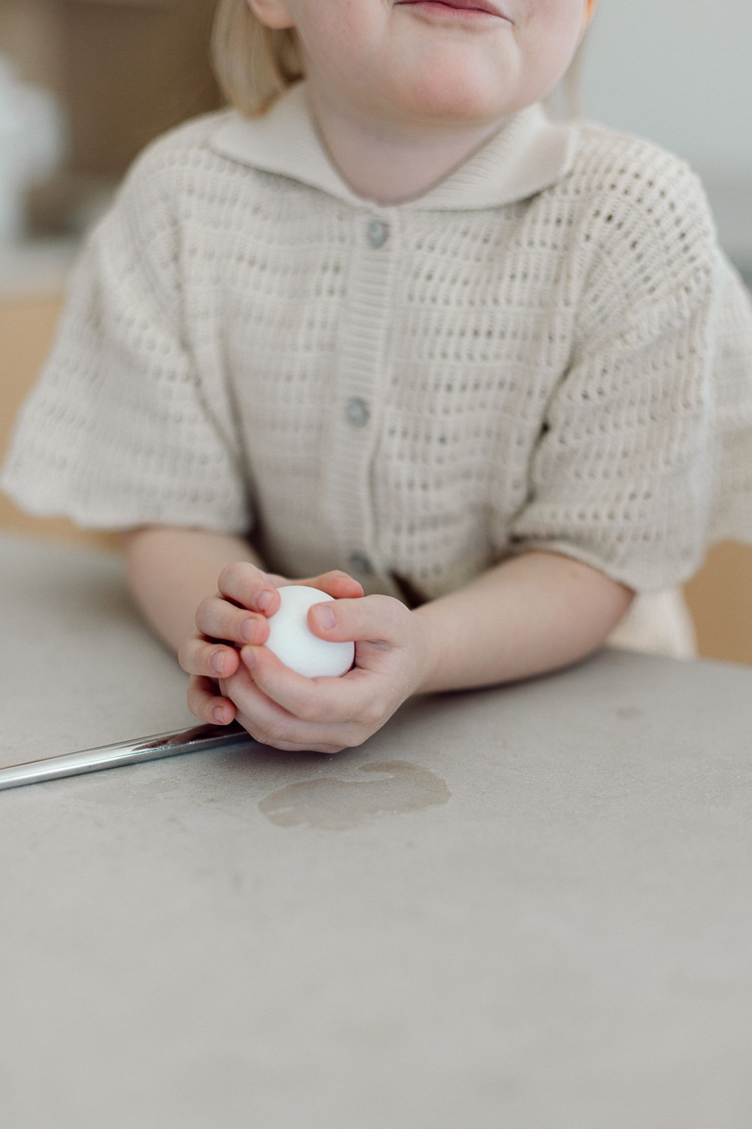 Young toddler in beige knit shirt holding a white egg at a table.