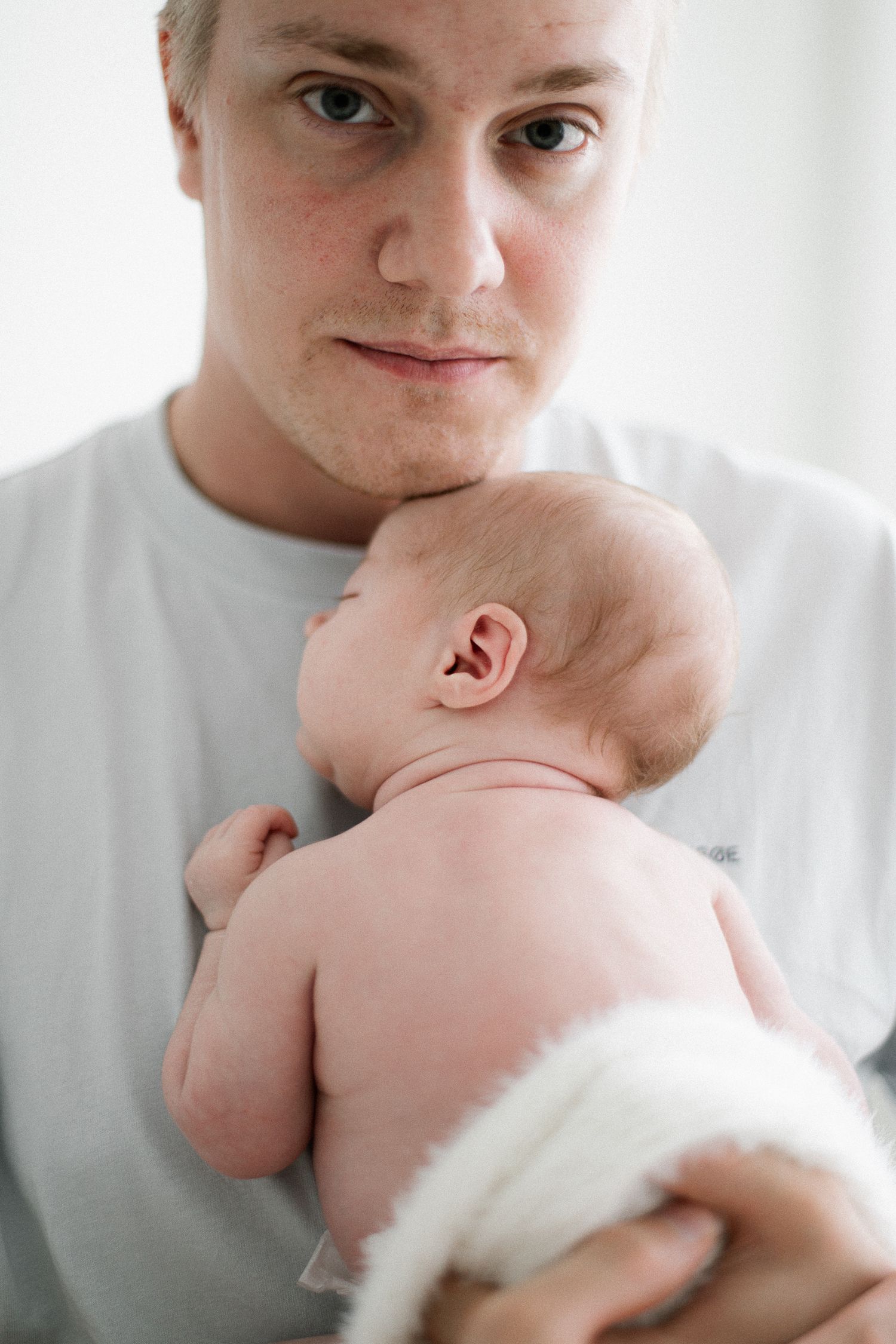 A young father in a gray shirt tenderly holds his newborn baby against his chest, looking at the camera.