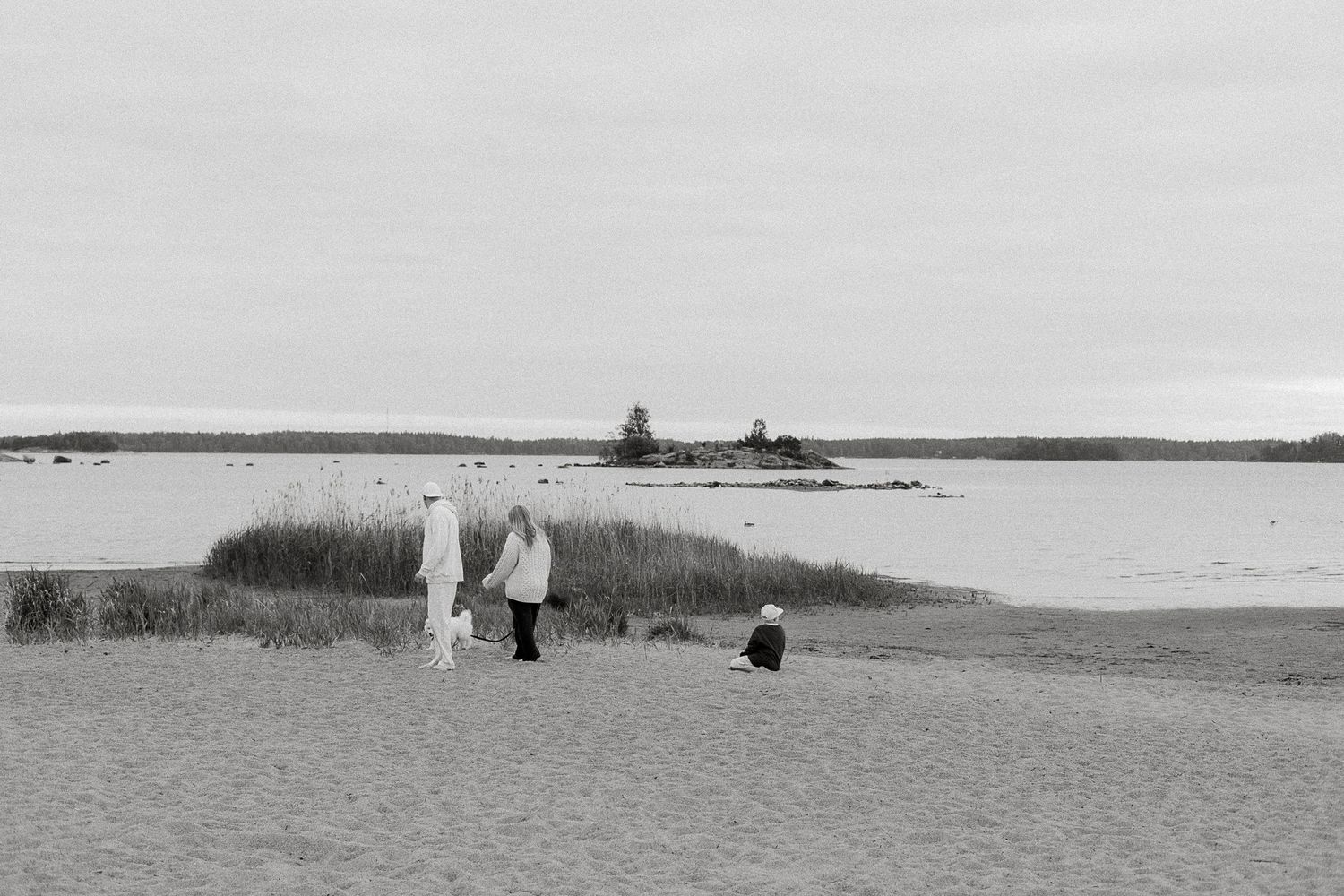 Black and white photo of people on a sandy beach with calm water and small island in the background.