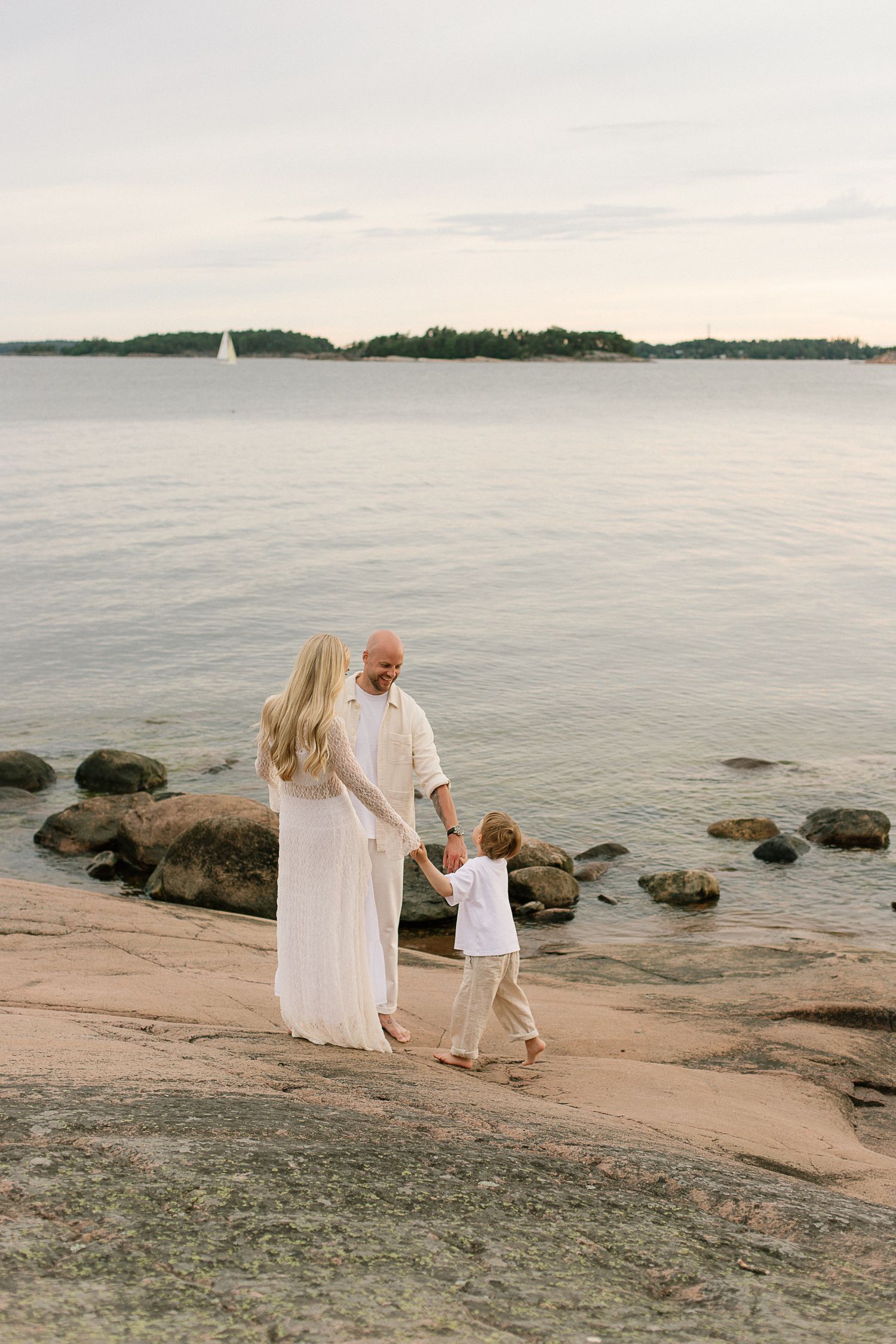 Family of three in white outfits walking on rocky shoreline by calm sea at dusk.
