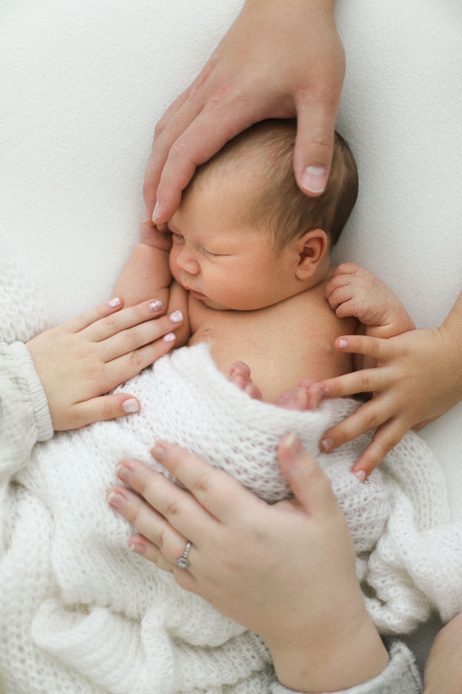 Newborn baby sleeping peacefully on white knit blanket while parents gently cradle with loving hands.